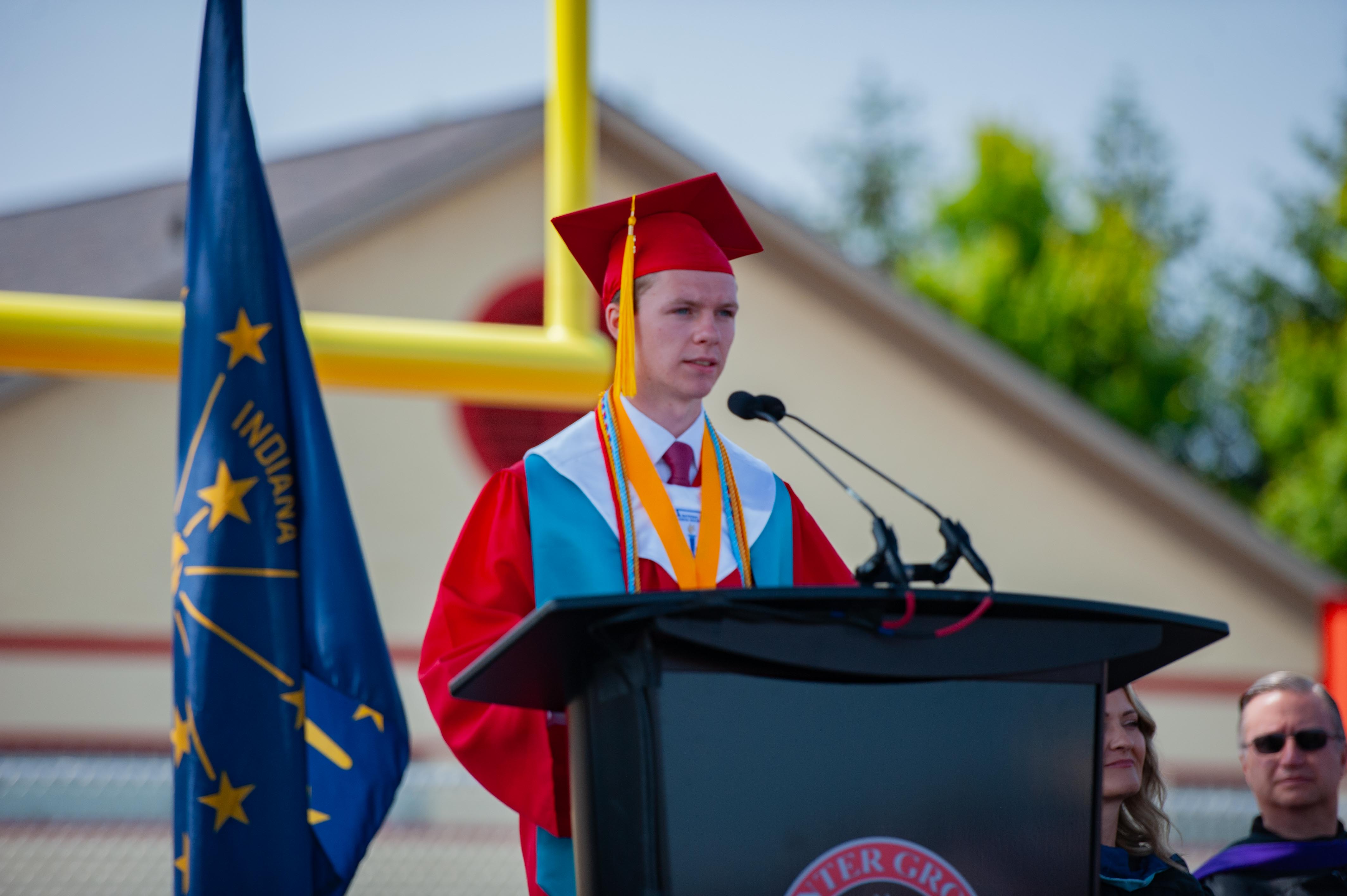 A young man in a graduation cap and gown speaks at a podium with an Indiana flag to his left and a yellow goalpost in the blurred background.
