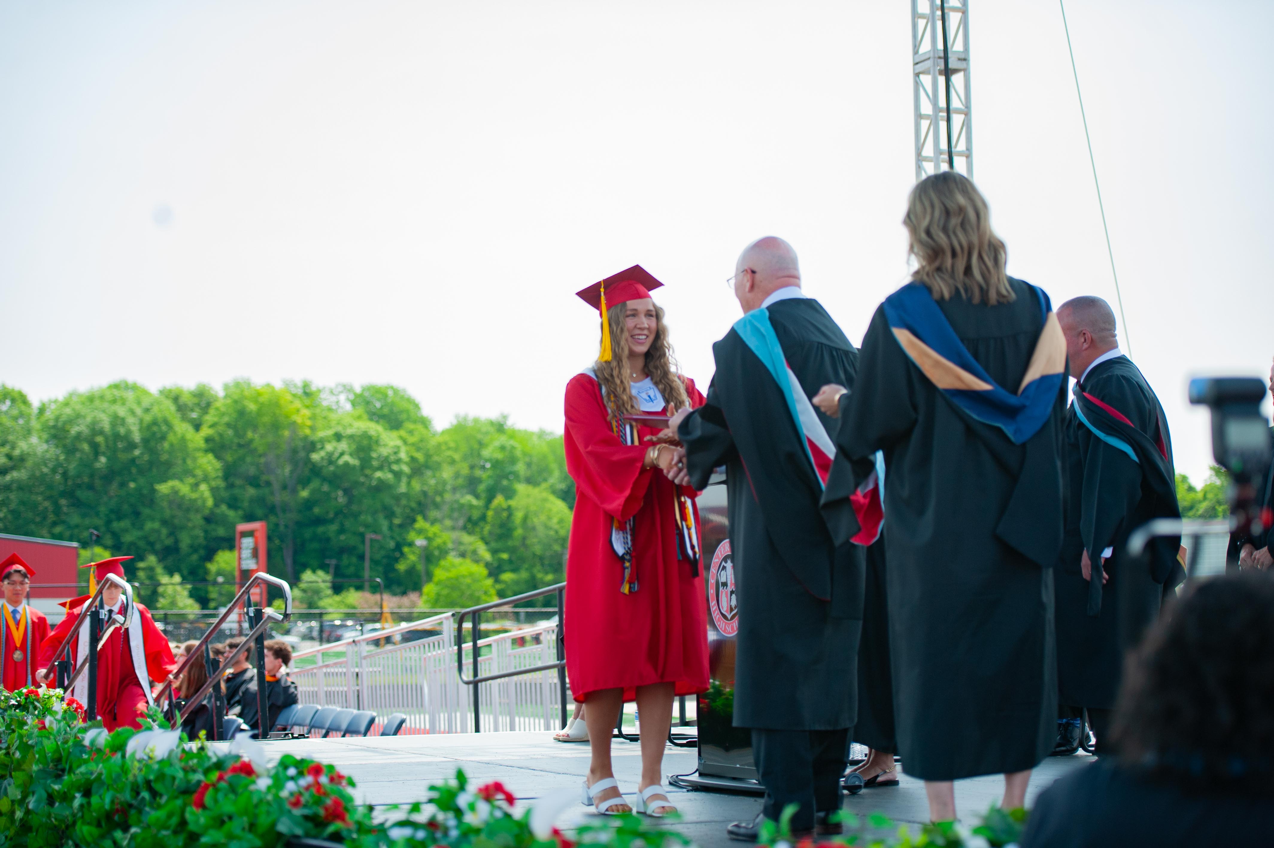 A graduating student in a red cap and gown receives a diploma on a stage with a lush green tree-lined background.