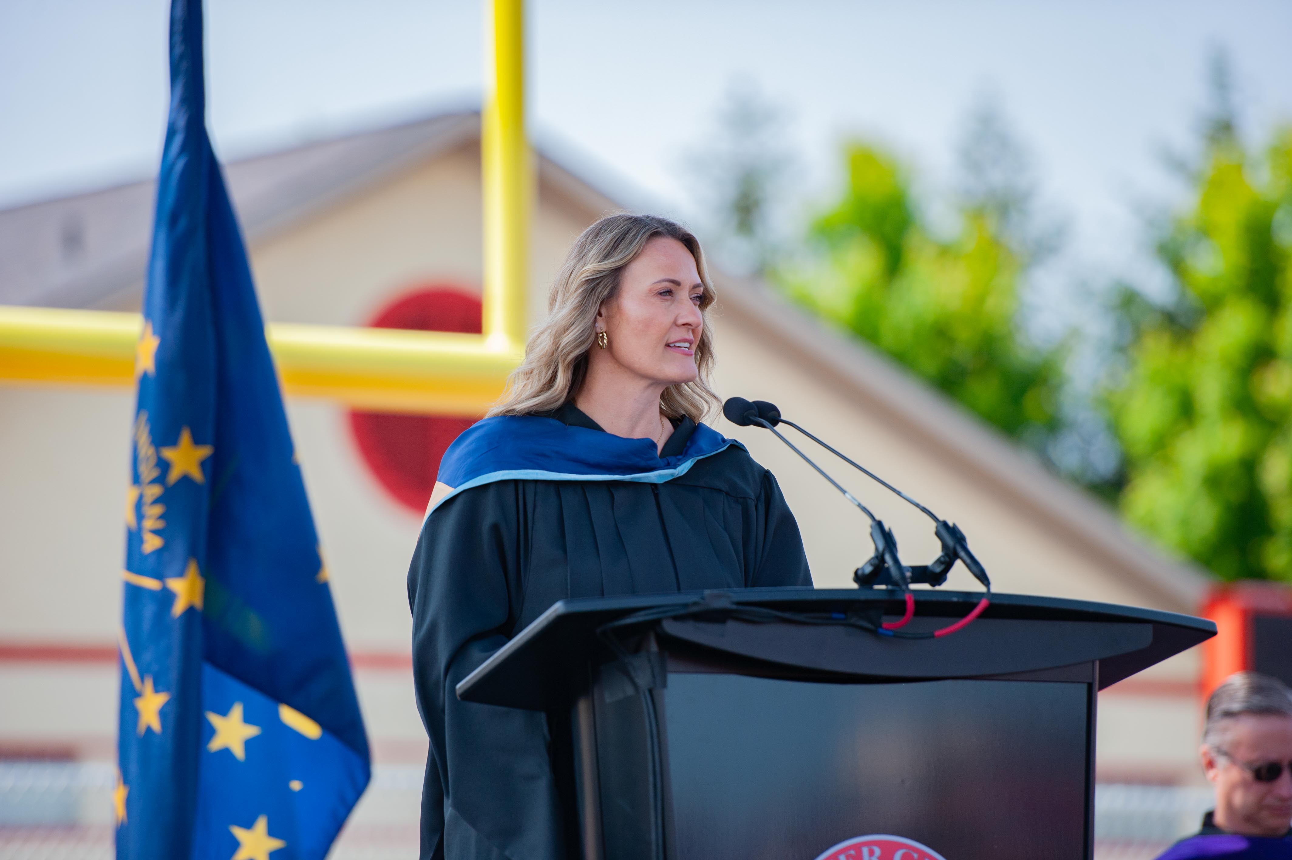 A woman in academic regalia speaks at a podium in front of a flag and a football goalpost.