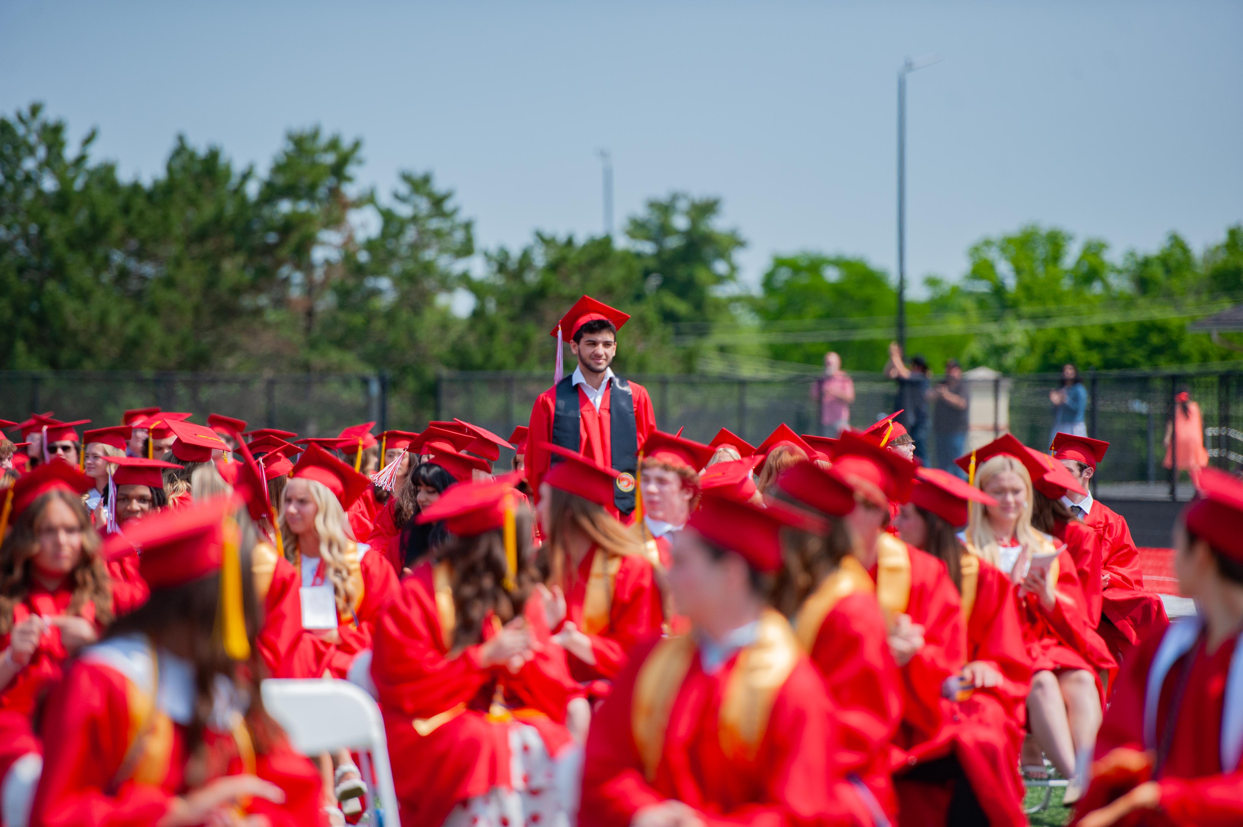 A young man in a graduation cap and gown stands above a crowd of graduates in red gowns and caps, with trees and a bright sky in the background.