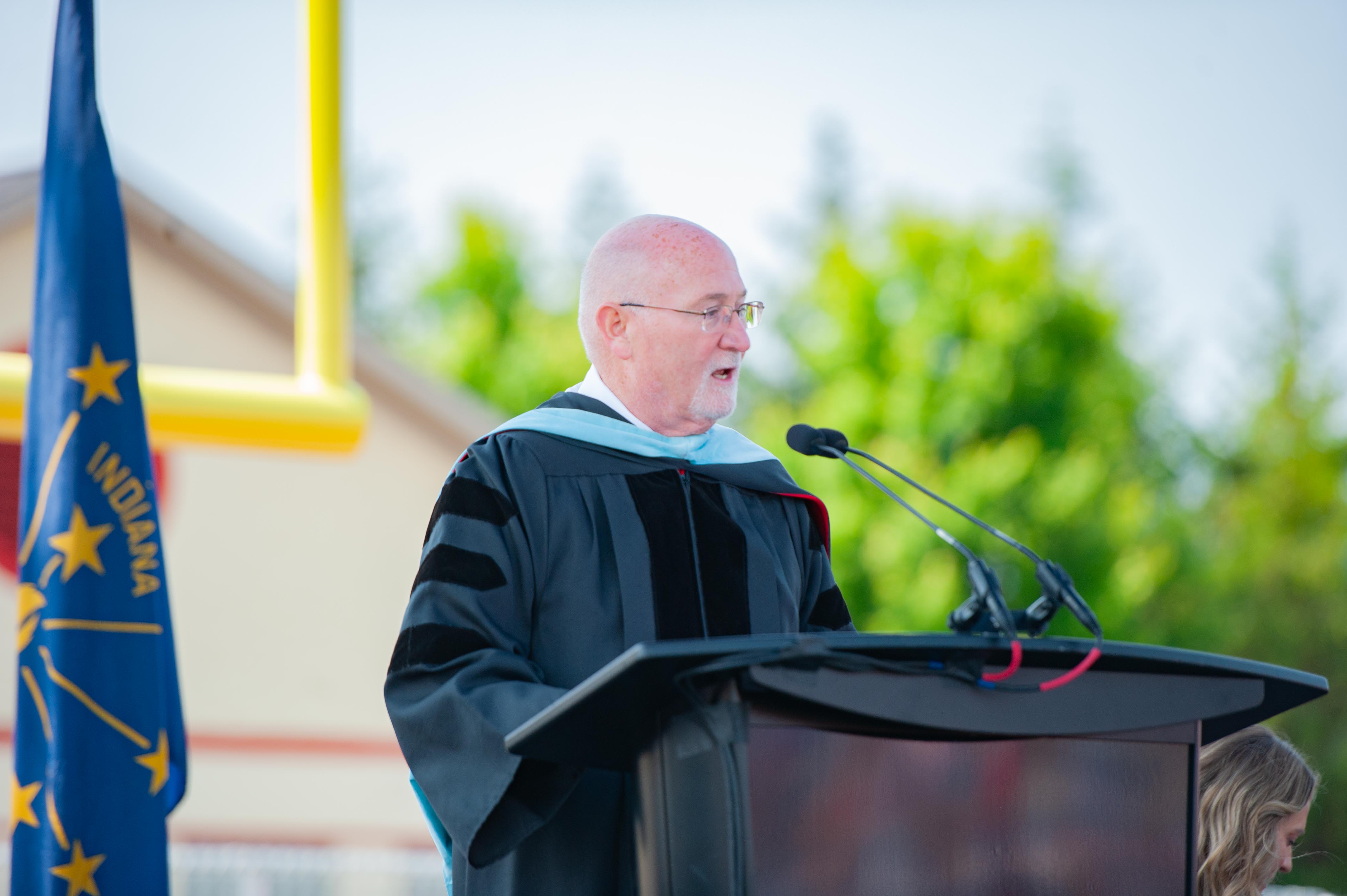 A bald man in academic regalia speaks at a podium with an Indiana flag to his left and blurred green trees and a yellow goalpost in the background.
