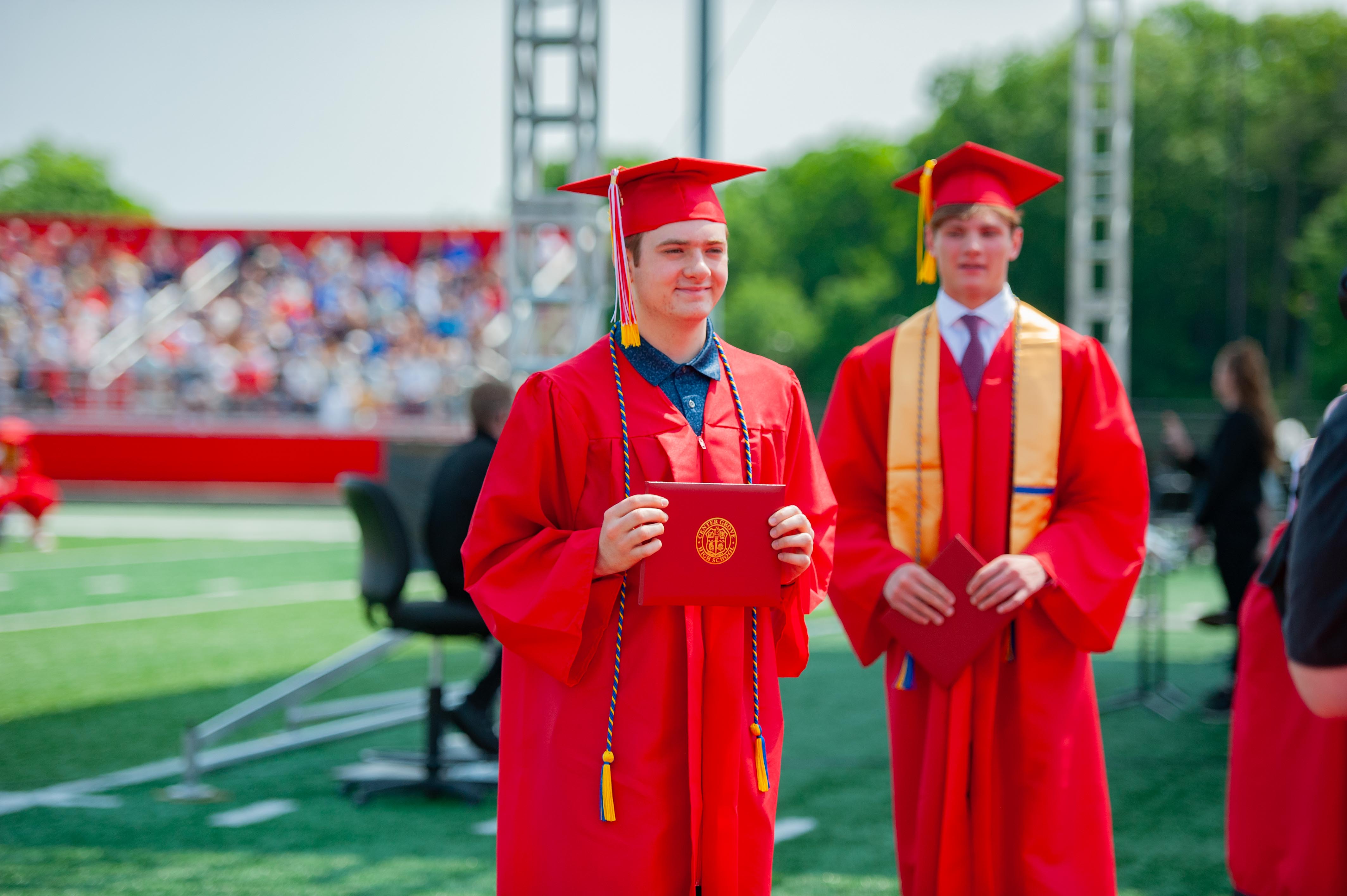 Two graduates in red gowns and caps stand on a green field, holding diplomas, with a blurred crowd in the background.