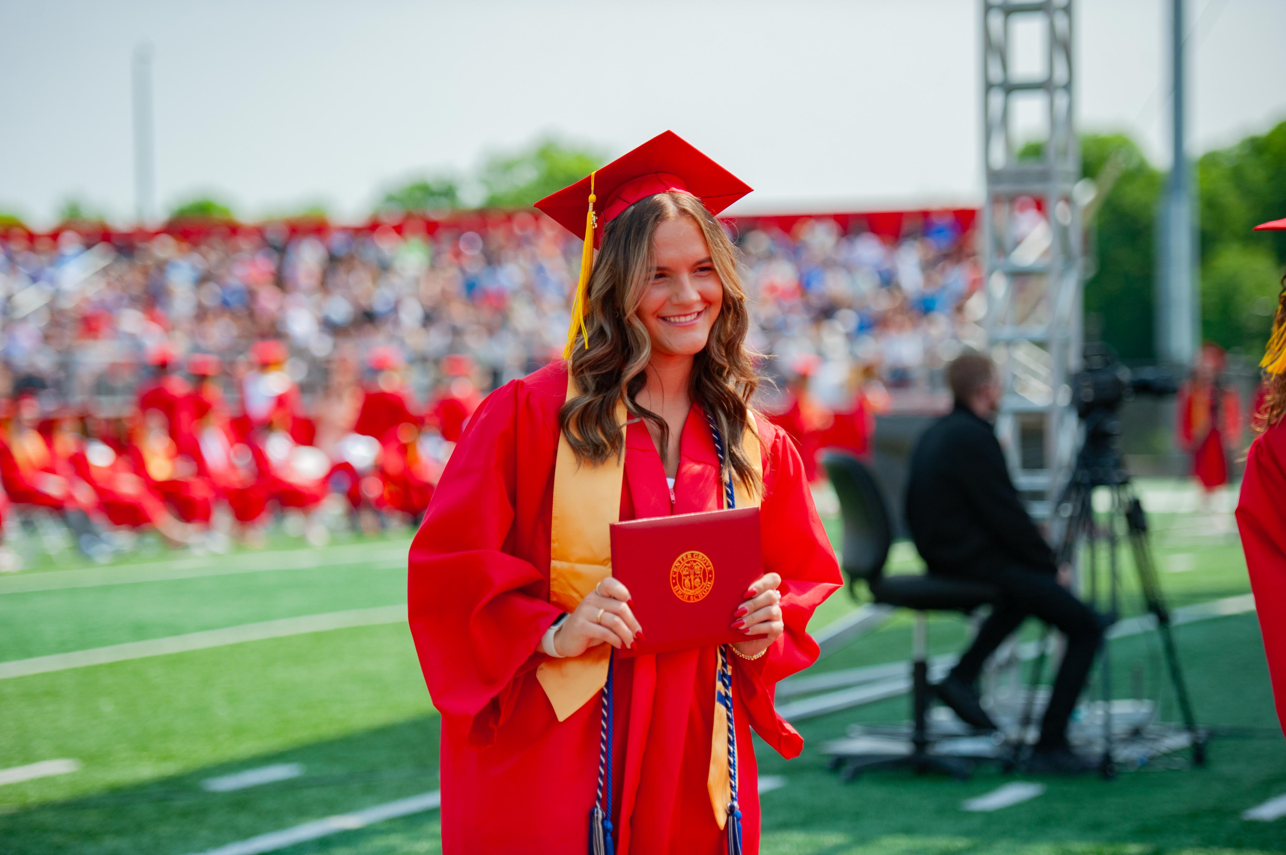 A smiling graduate in a red cap and gown holds a diploma on a green field with a blurred crowd in the background.