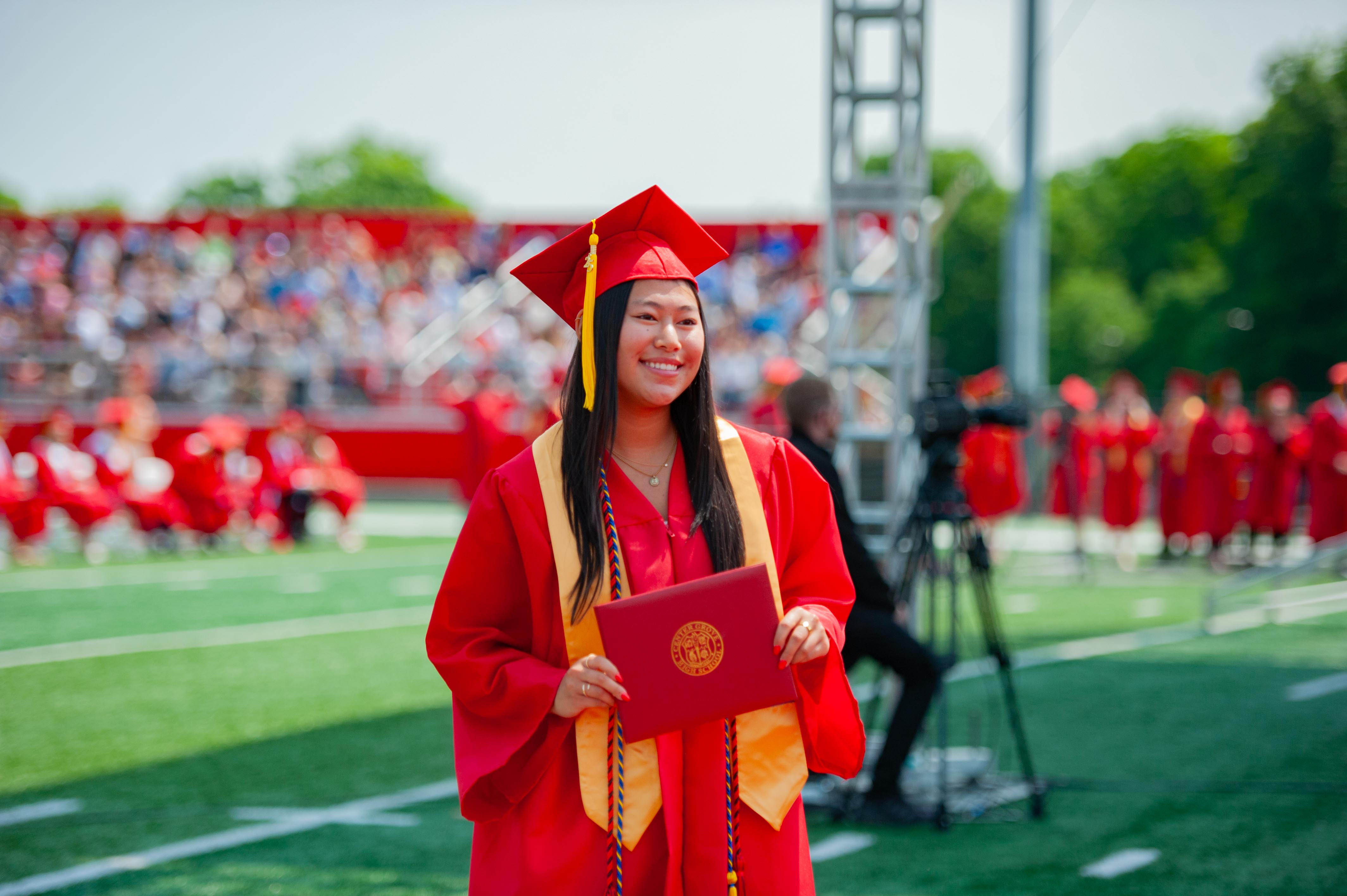 A smiling graduate in a red cap and gown holds a diploma on a green field with a blurred crowd in the background.