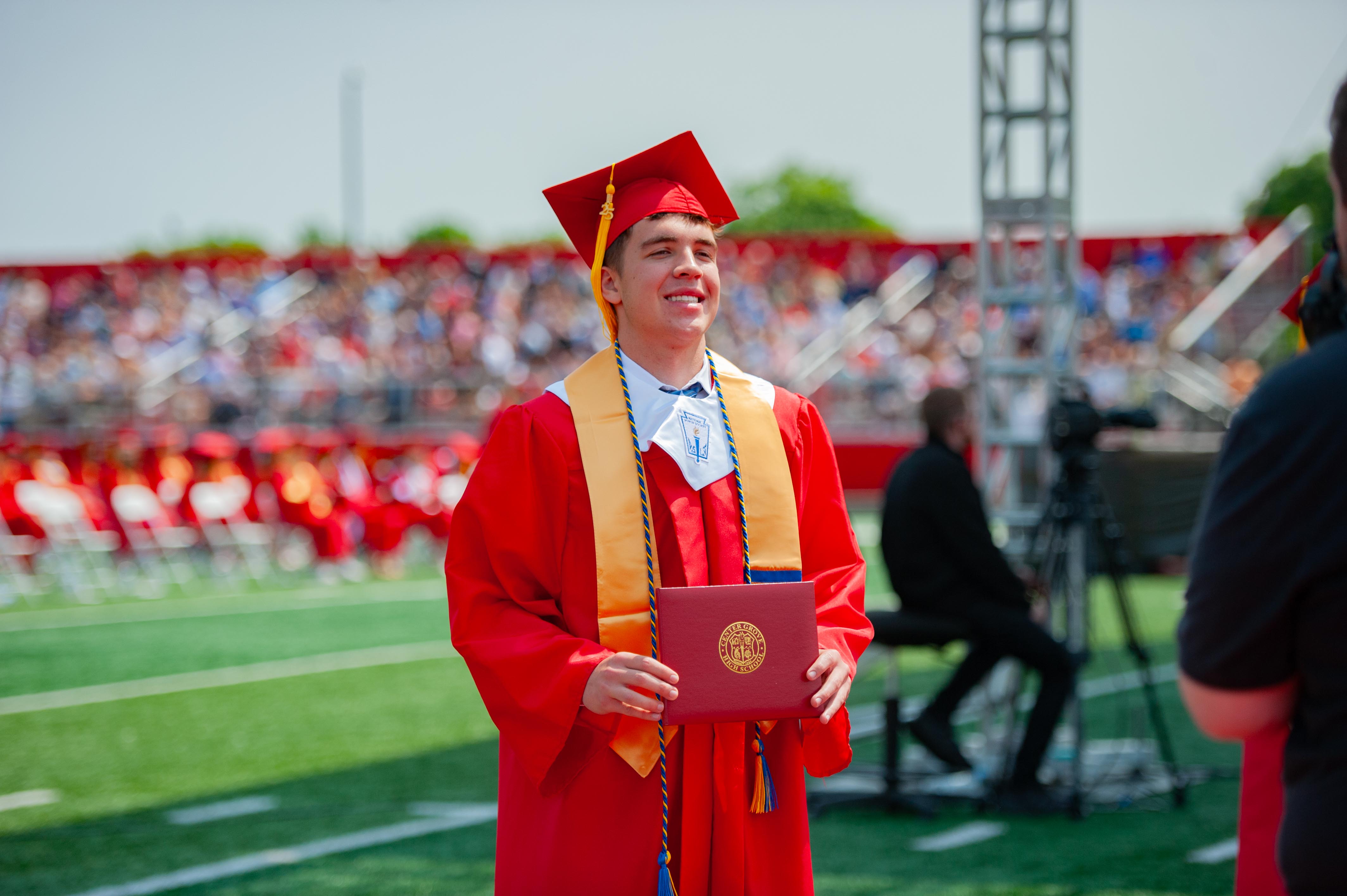 A smiling graduate in a red cap and gown holds a diploma on a green field with a blurred audience in the background.