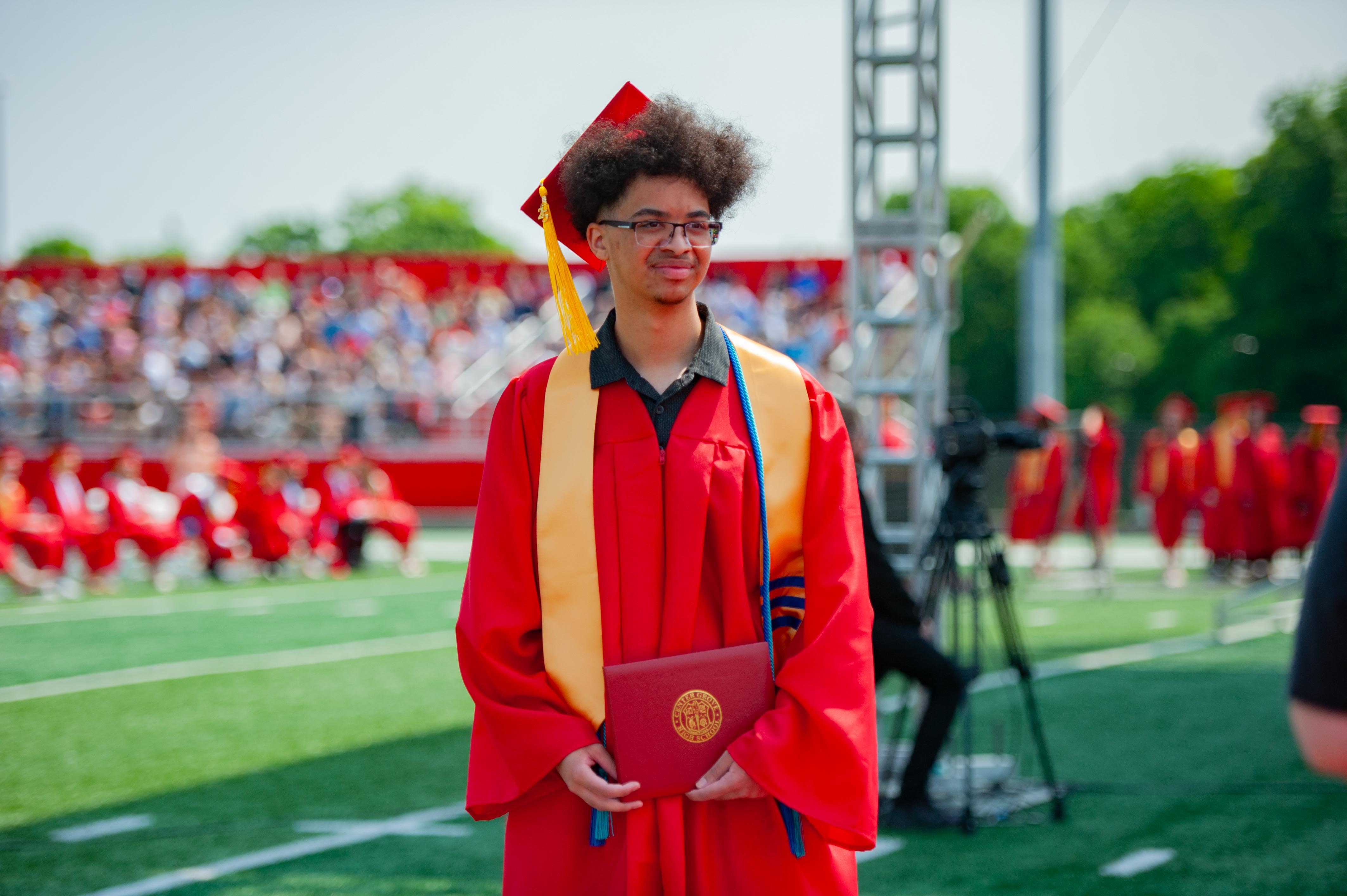 A young man in a red graduation gown and cap stands on a green field holding a diploma, with a crowd of graduates and spectators in the blurred background.