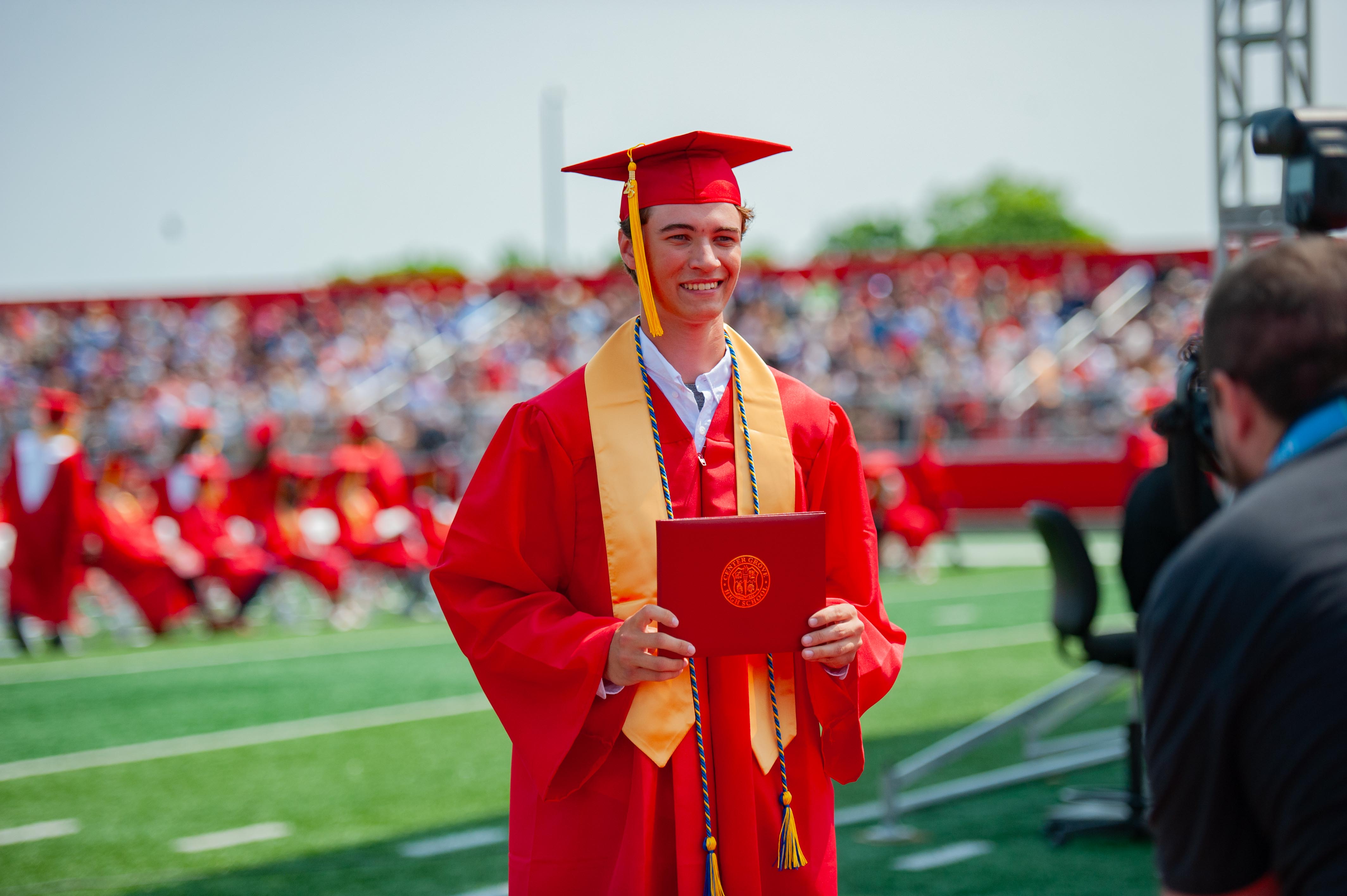 A smiling graduate in a red cap and gown holds a diploma on a green football field with a blurred crowd in red bleachers in the background.