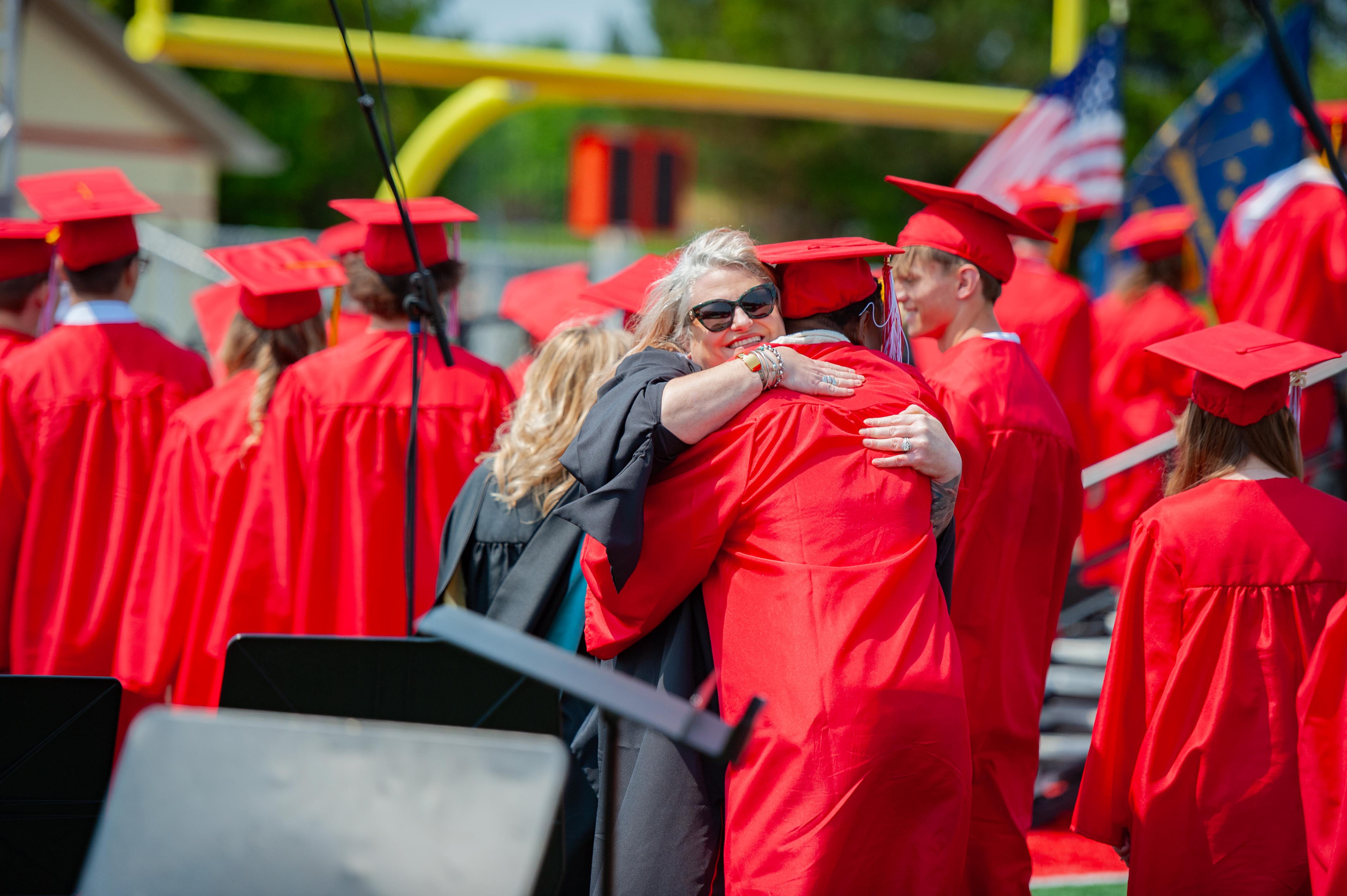 A woman in a black robe hugs a graduate in a red cap and gown, while other graduates in red attire stand in the background with flags and a football goalpost.
