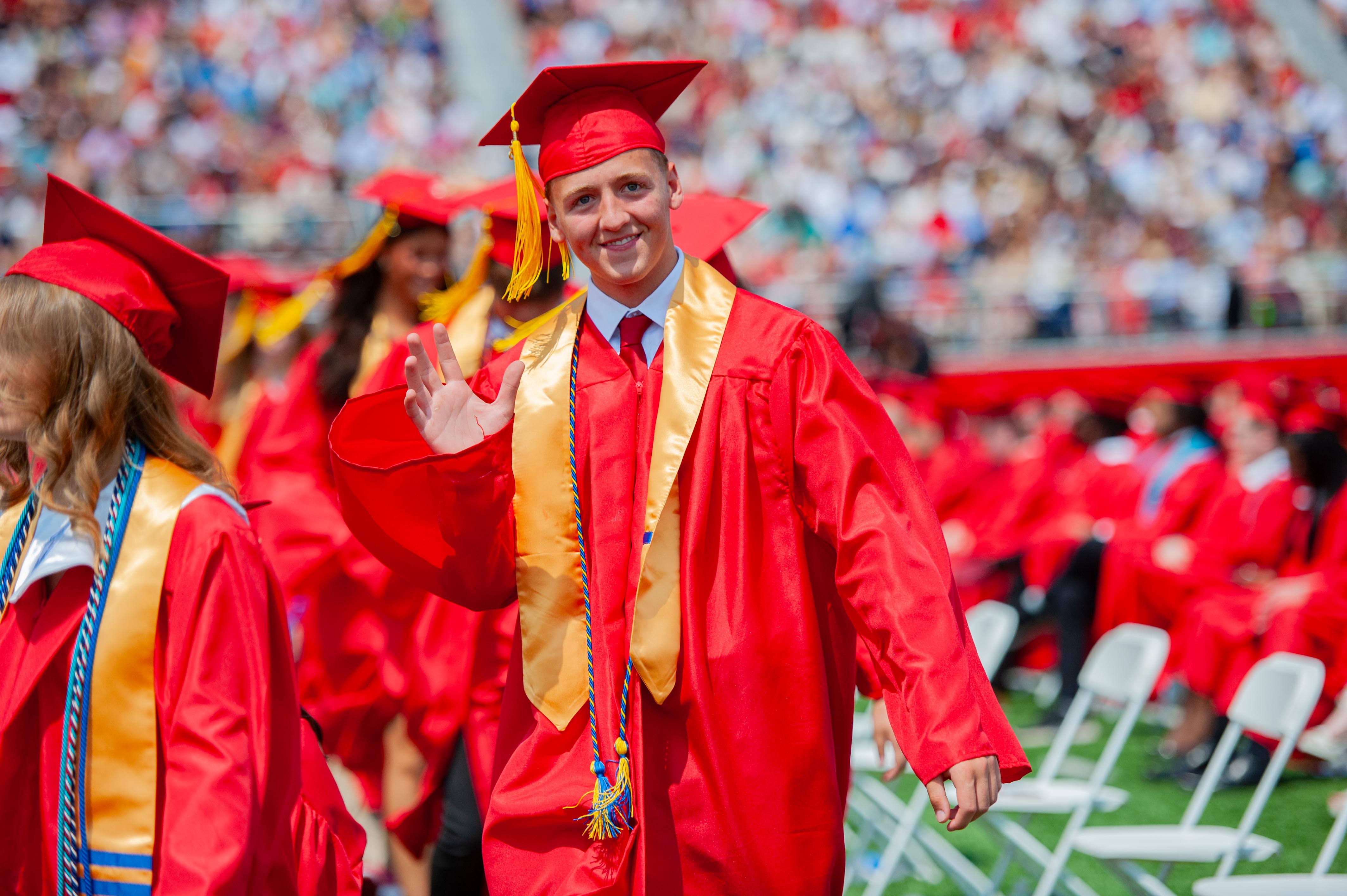 A smiling graduate in a red cap and gown waves to the crowd in the background during a graduation ceremony.