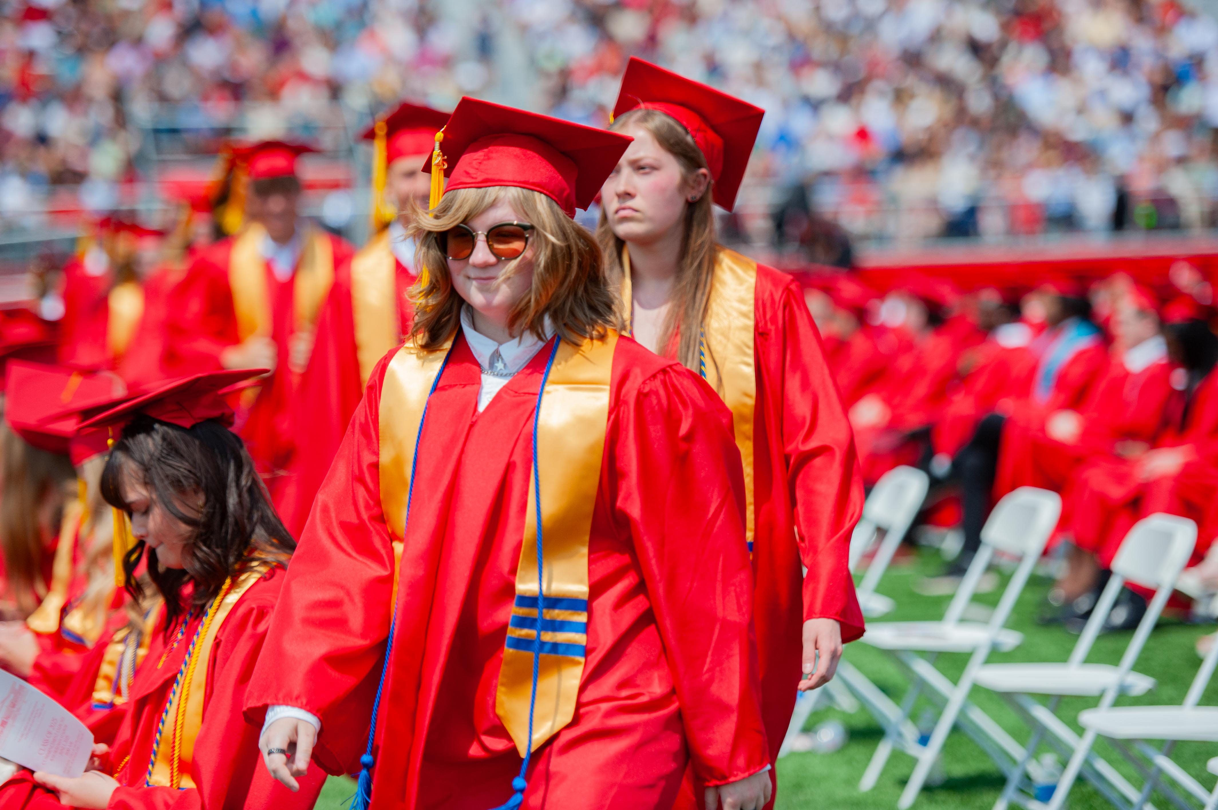 Graduates in red caps and gowns walk across a field with a blurred audience in the background.