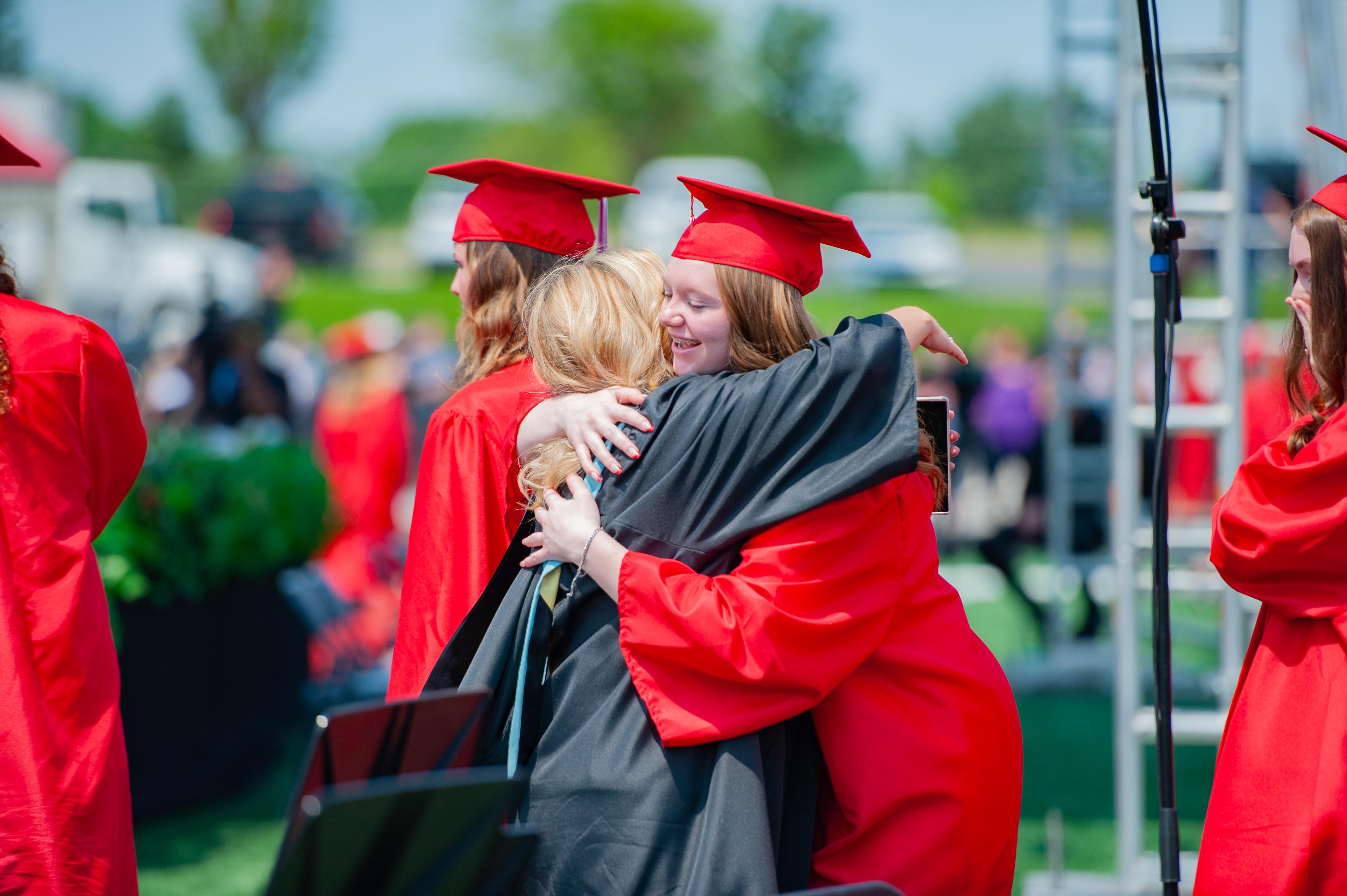 Two graduates in red caps and gowns embrace in the foreground, while a blurred crowd and white tents are visible in the background under a bright sky.