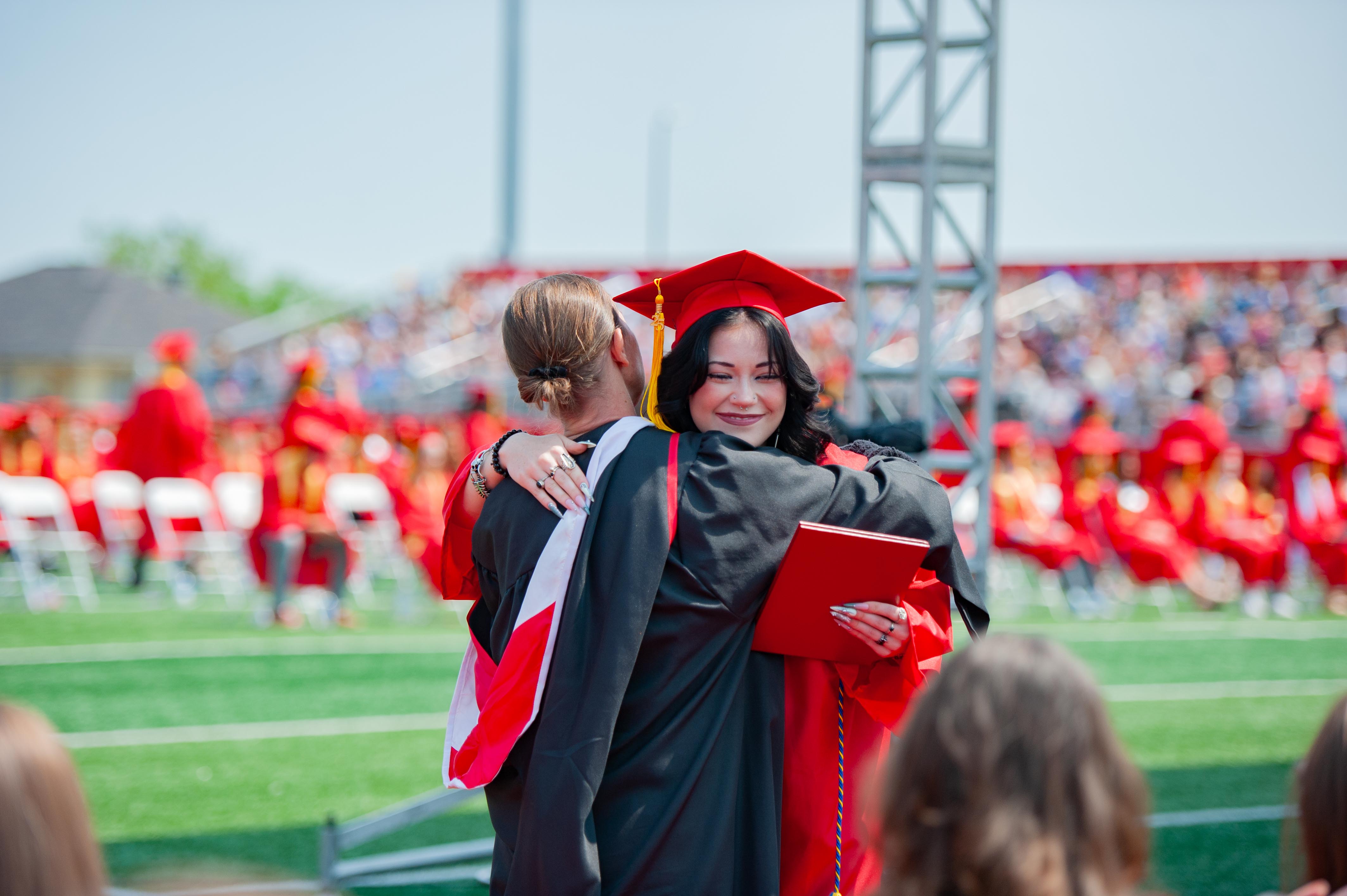 Two graduates in red caps and gowns embrace on a green field, with a blurred crowd of spectators in the background.