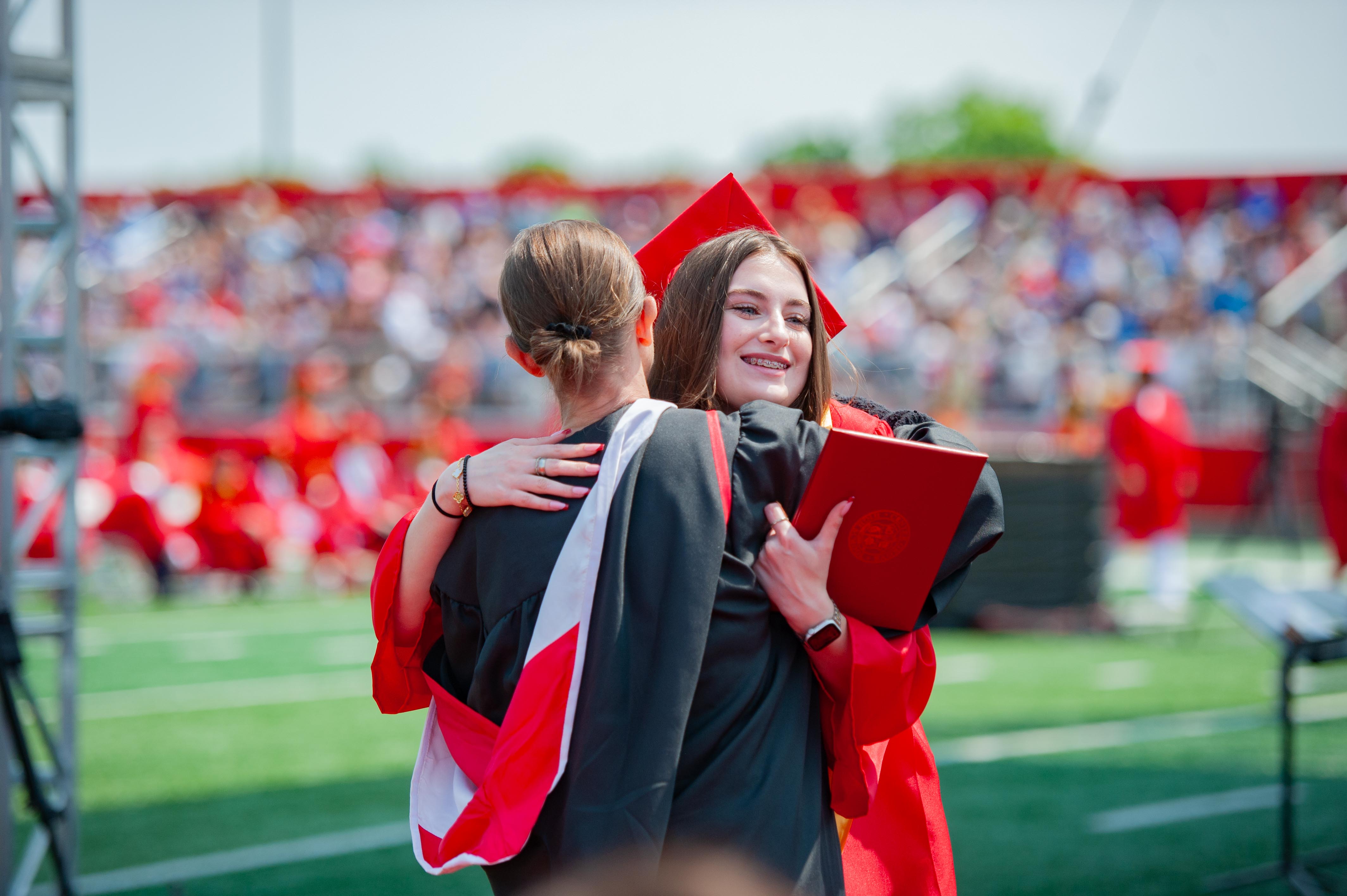 Two graduates in red and black gowns embrace on a green field with a blurred crowd in red attire in the background.