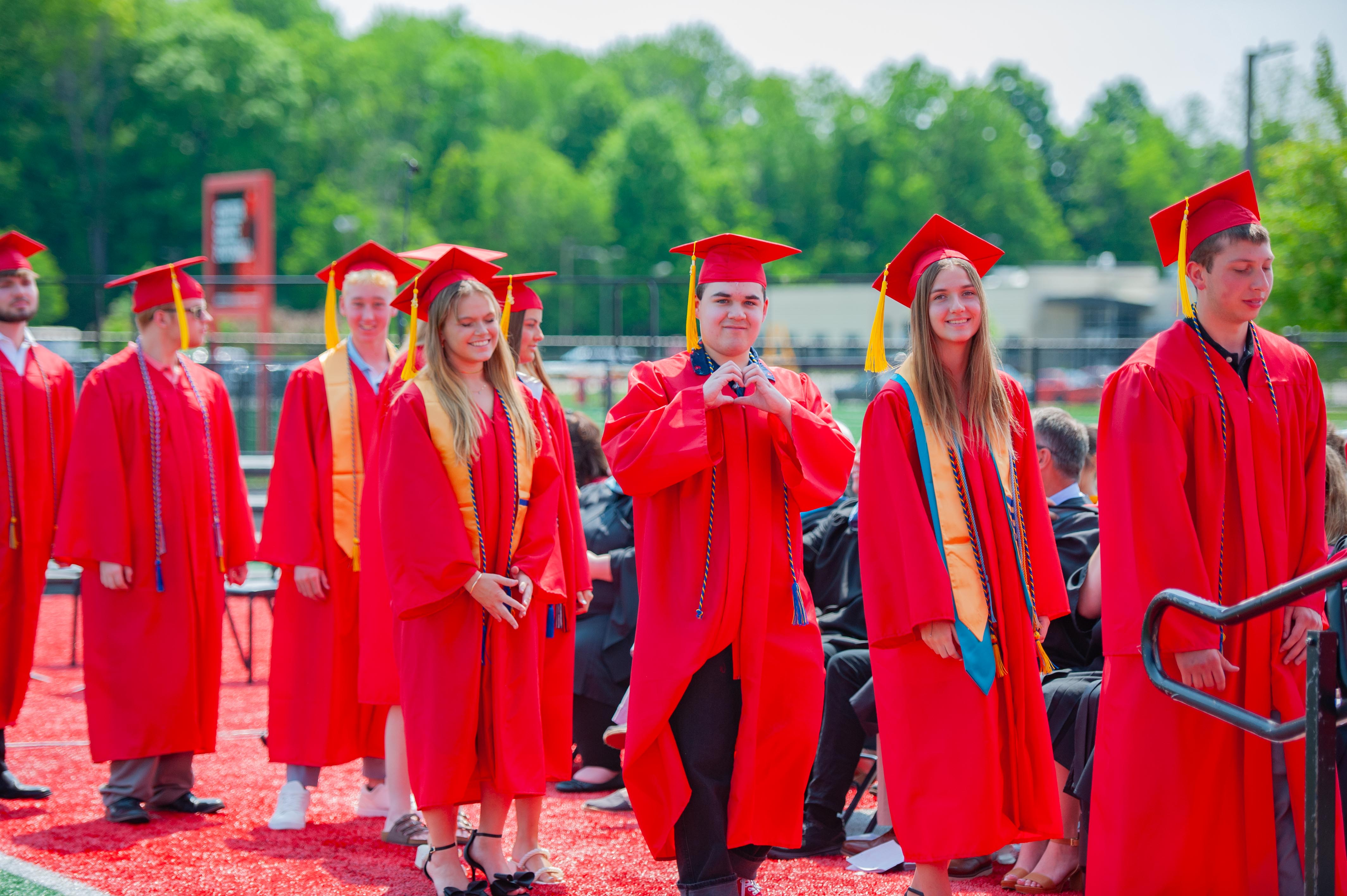 A group of graduates in red gowns and caps walk in a line on a grassy field with trees and buildings in the background.