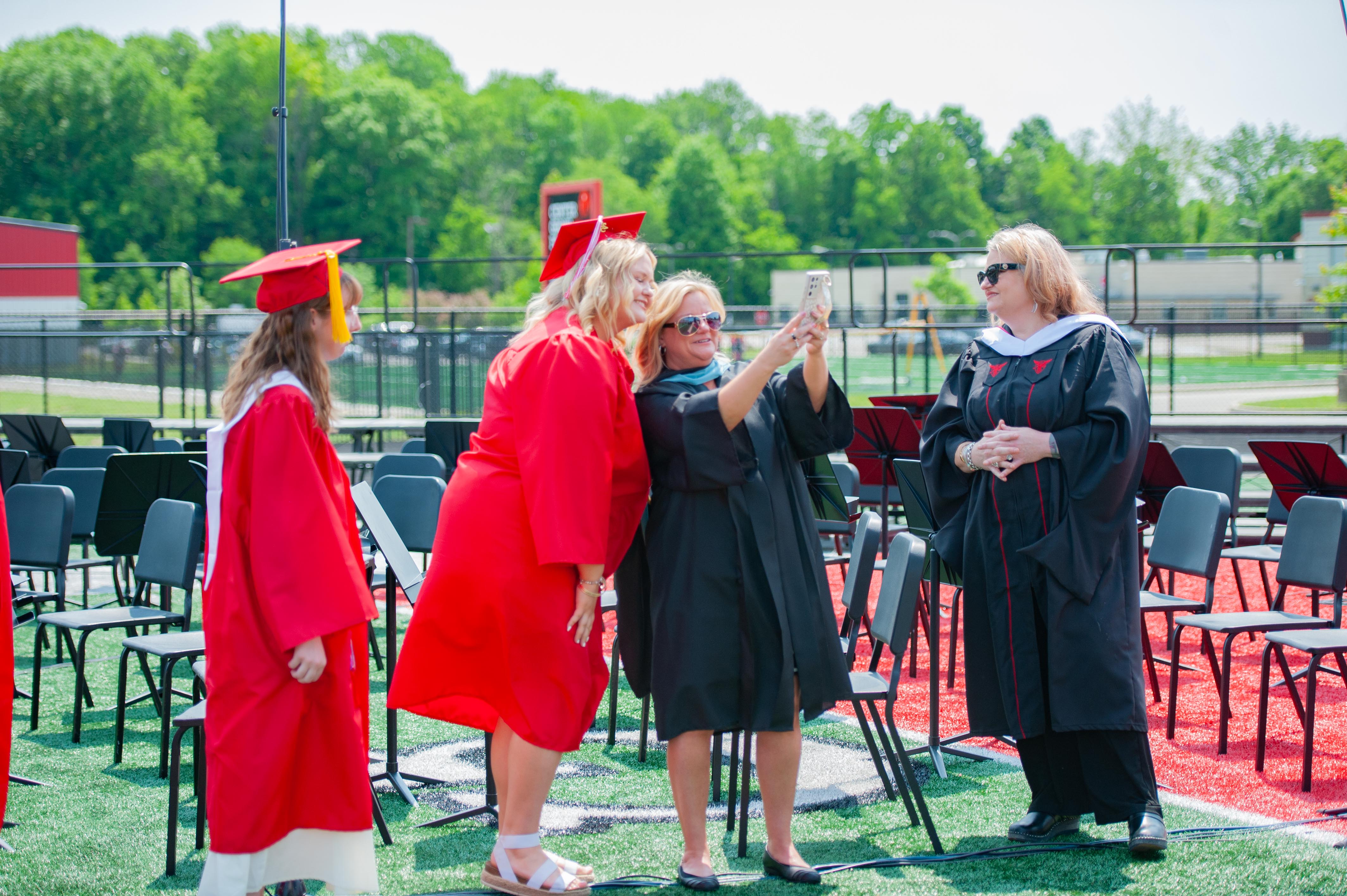 Four graduates in red gowns and caps pose for a selfie in the foreground, with rows of empty chairs and a grassy field in the background.