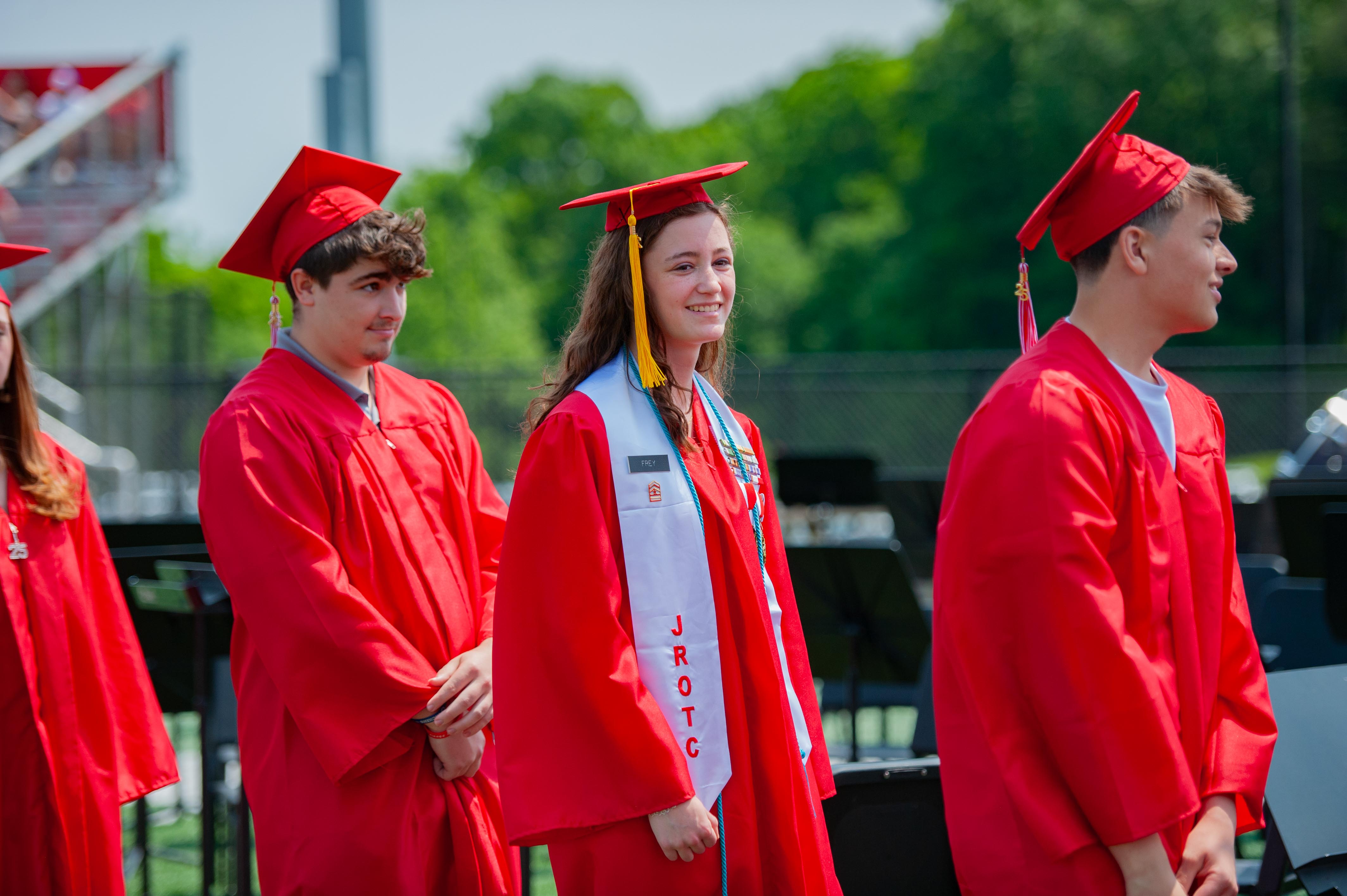 Graduating students in red caps and gowns stand in a line on a field with bleachers and trees in the background.