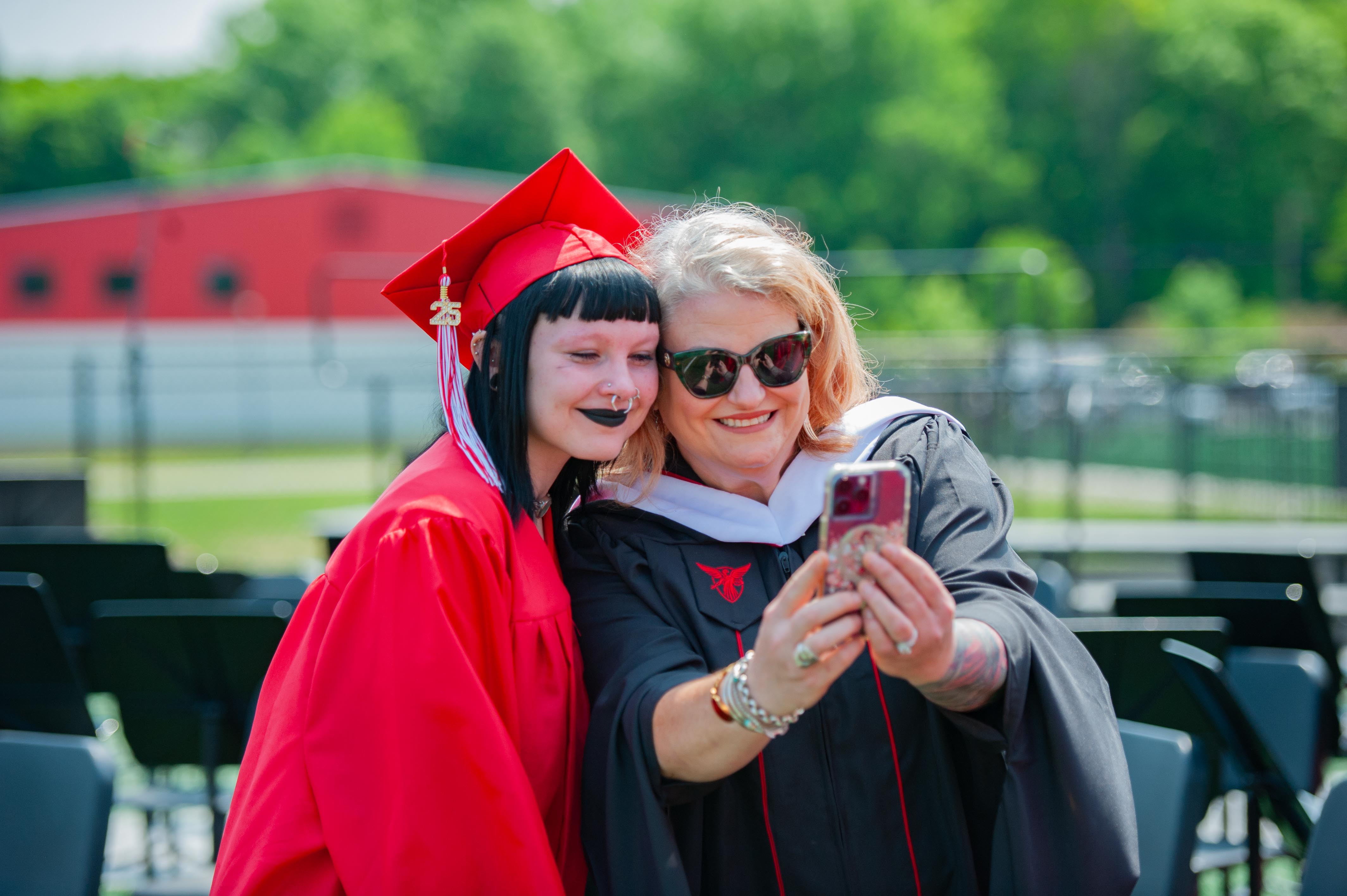 A graduate in a red cap and gown poses for a selfie with a woman in academic regalia, with a red building and green trees in the background.