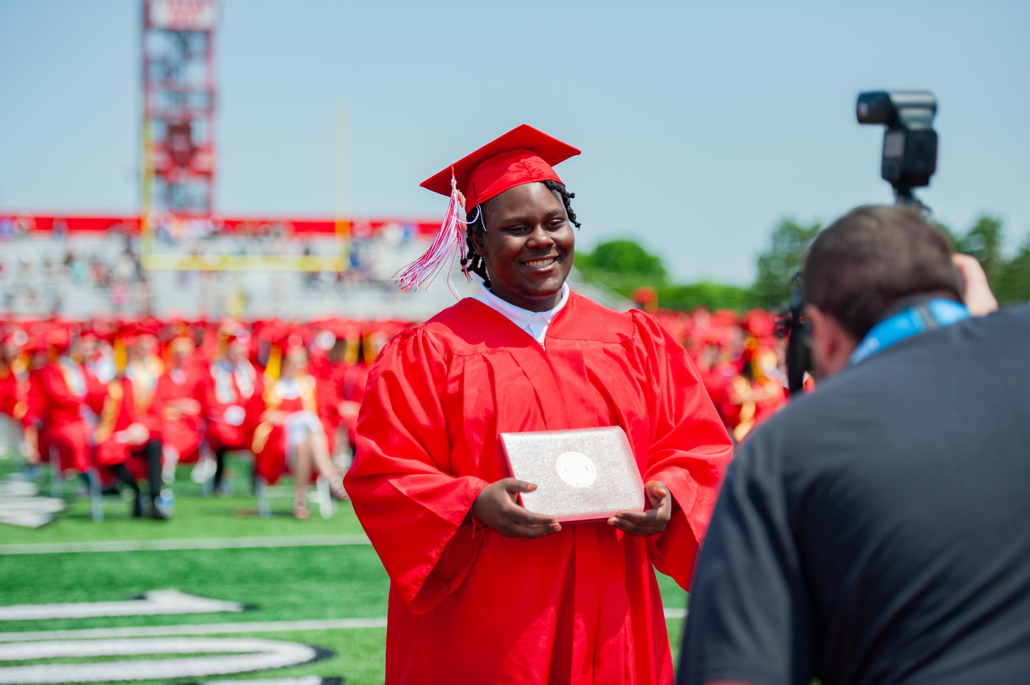 A graduate in a red cap and gown smiles while holding a diploma on a football field, with a photographer in the foreground and a stadium filled with spectators in the background.