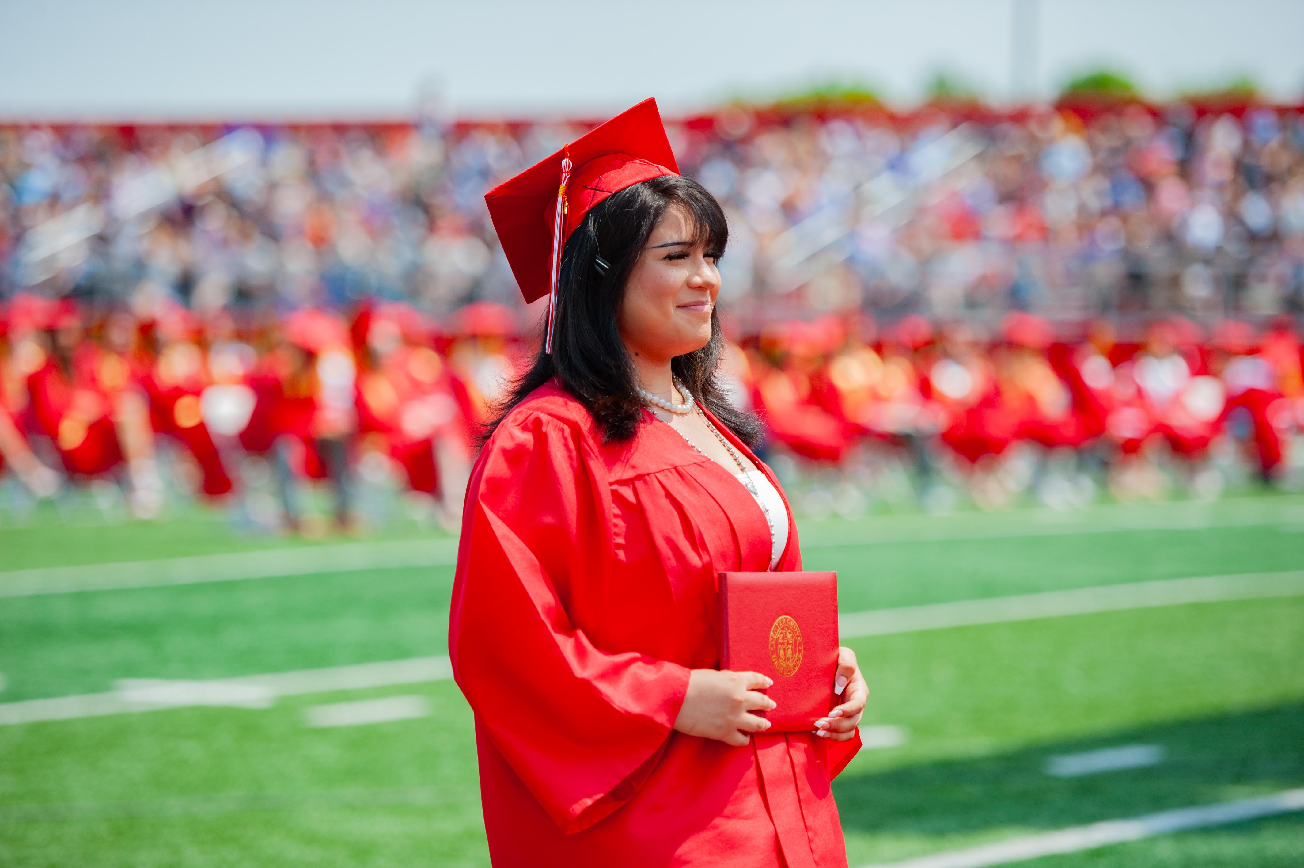 A smiling graduate in a red cap and gown holds a diploma on a green football field with a blurred crowd in the background.