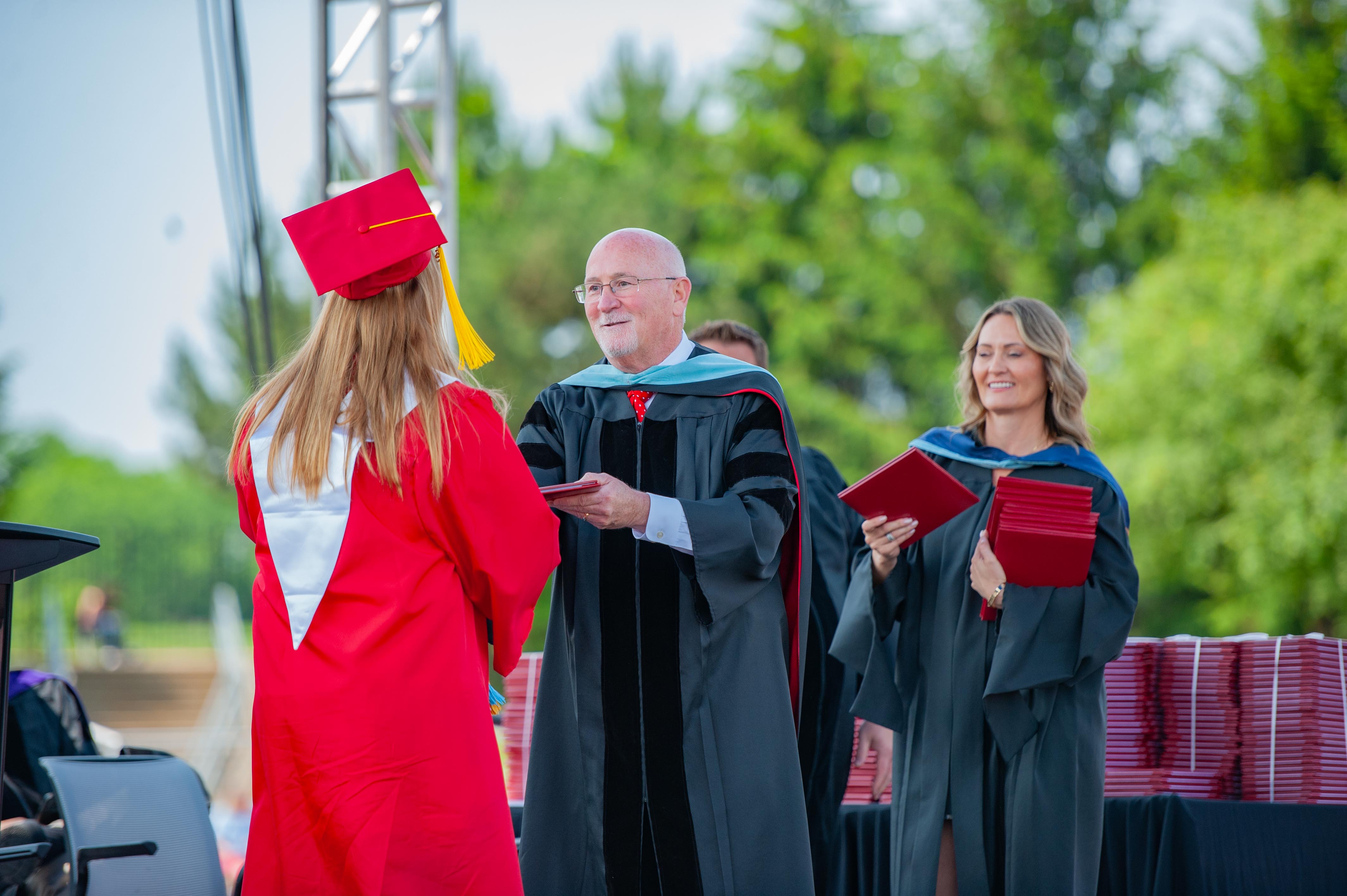 A graduate in a red cap and gown receives a diploma from an administrator, with a smiling woman in academic regalia holding more diplomas in the background against a backdrop of trees.