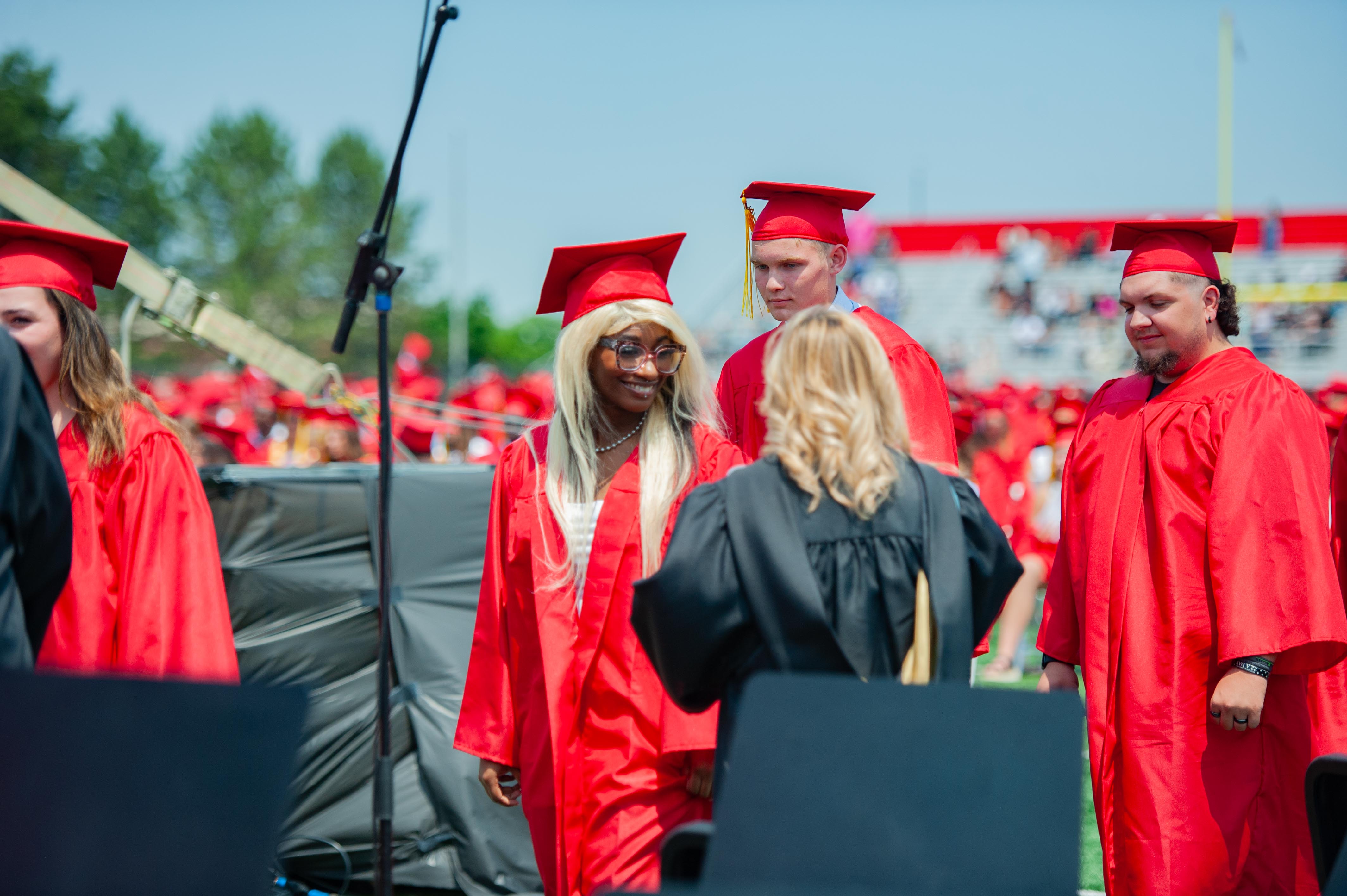 Graduates in red gowns and caps are walking across a stage with a blurred audience in the background.