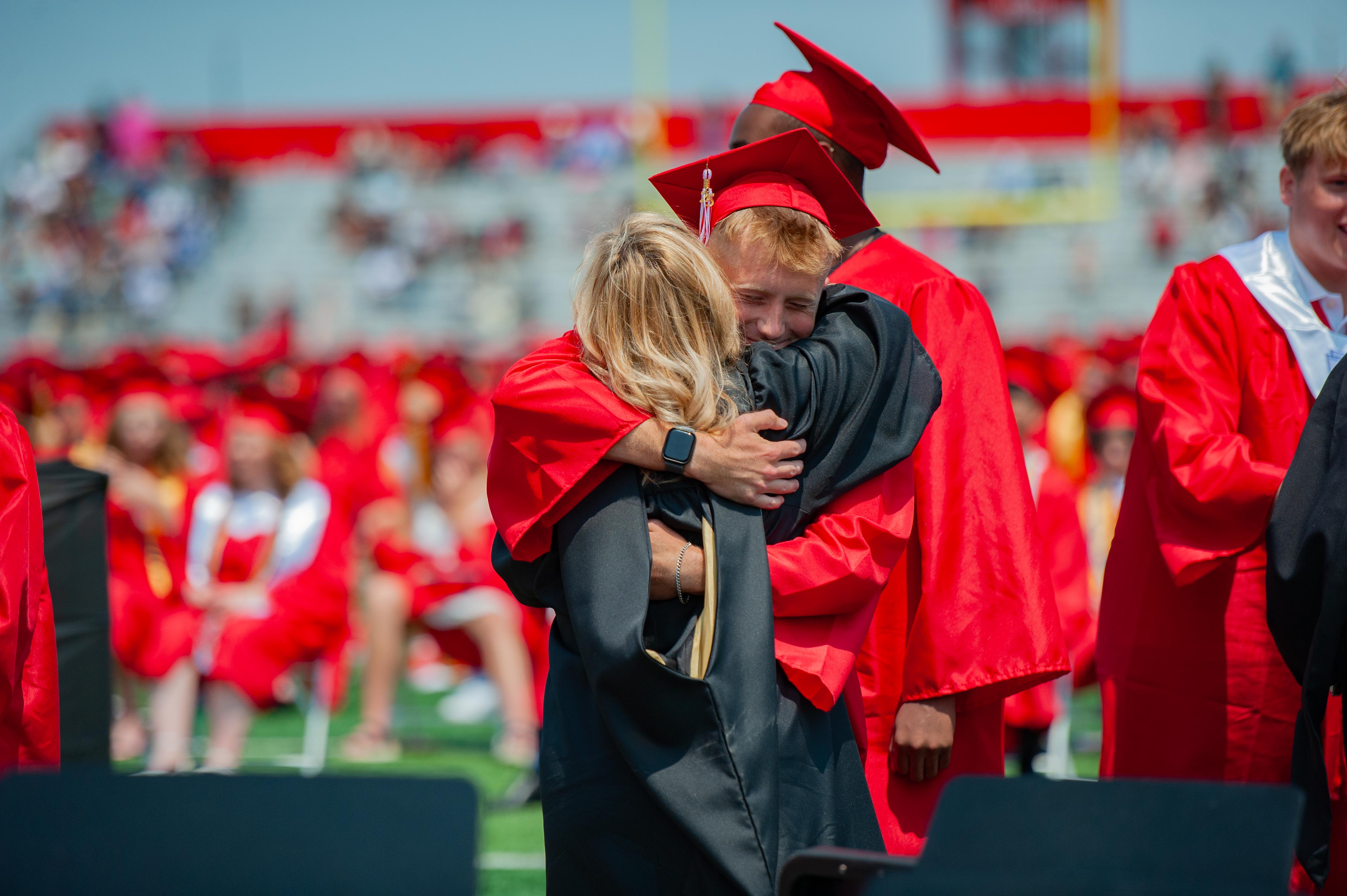 A graduate in a black gown and red cap is embraced by a woman in red, with a stadium full of people in red caps and gowns in the background.