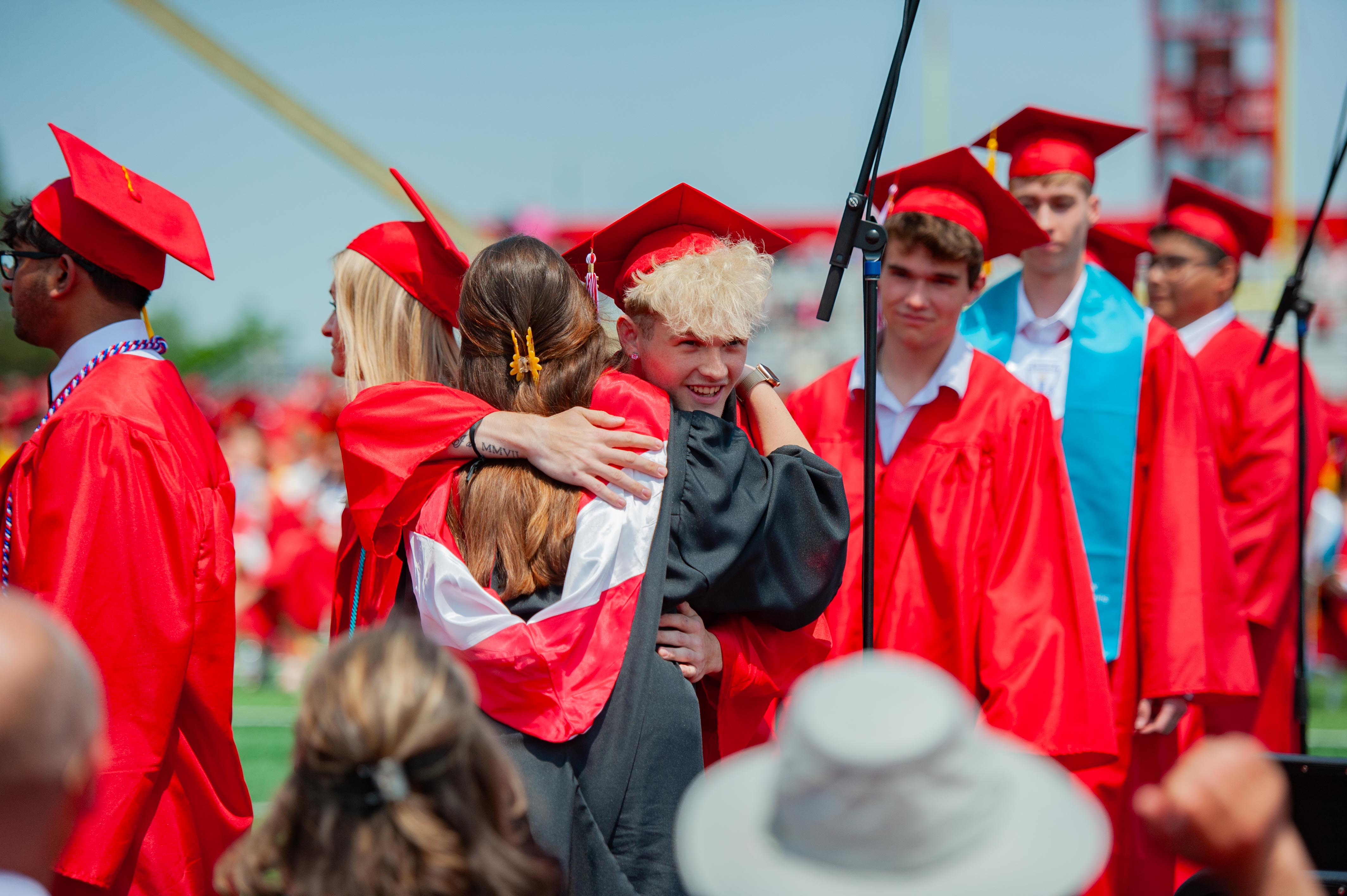 Graduates in red gowns and caps embrace in the foreground, while a crowd and stadium bleachers are visible in the background.