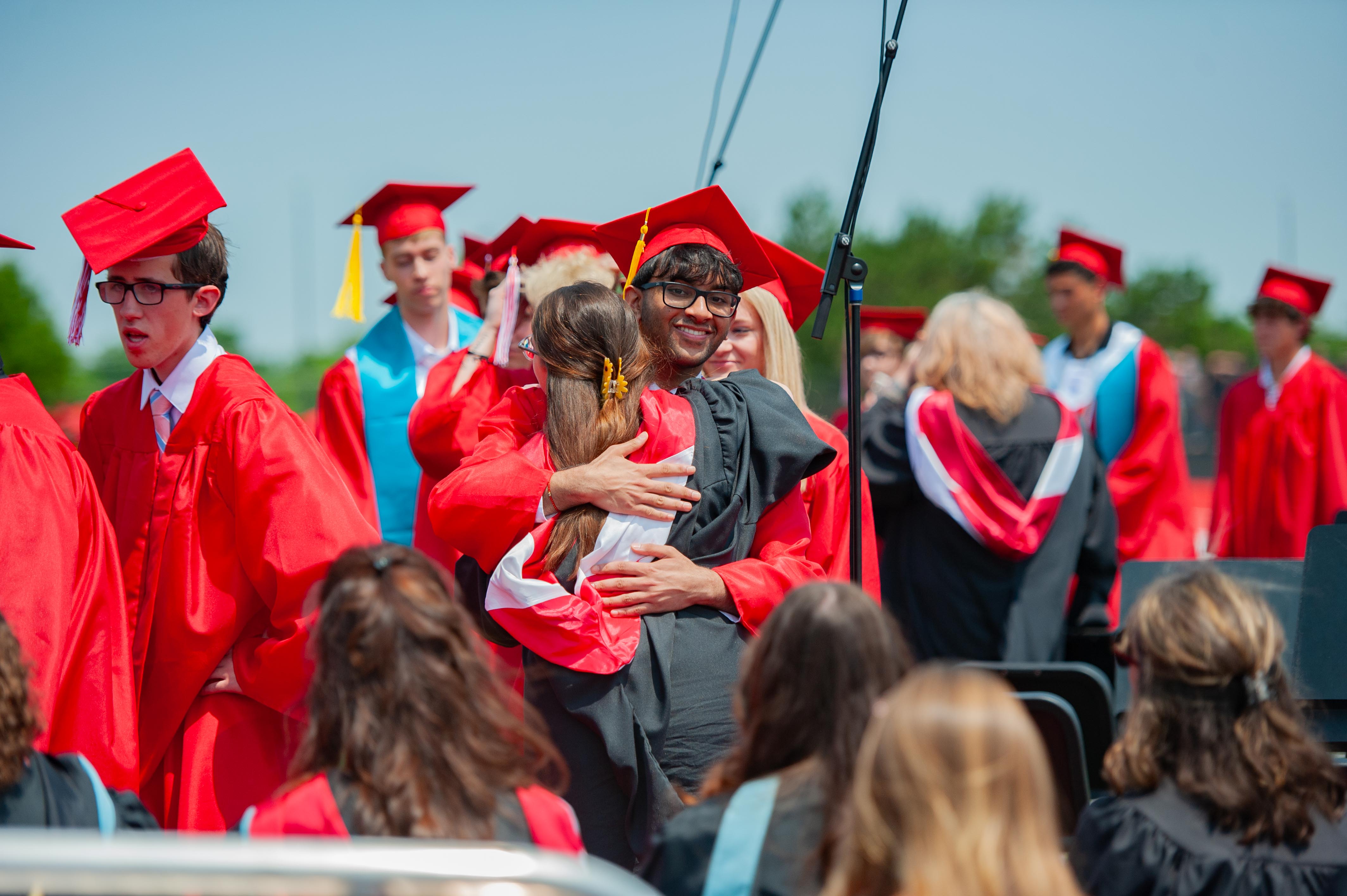 In the foreground, graduates in red gowns embrace, while in the background, more graduates in red and blue gowns stand under a bright sky.