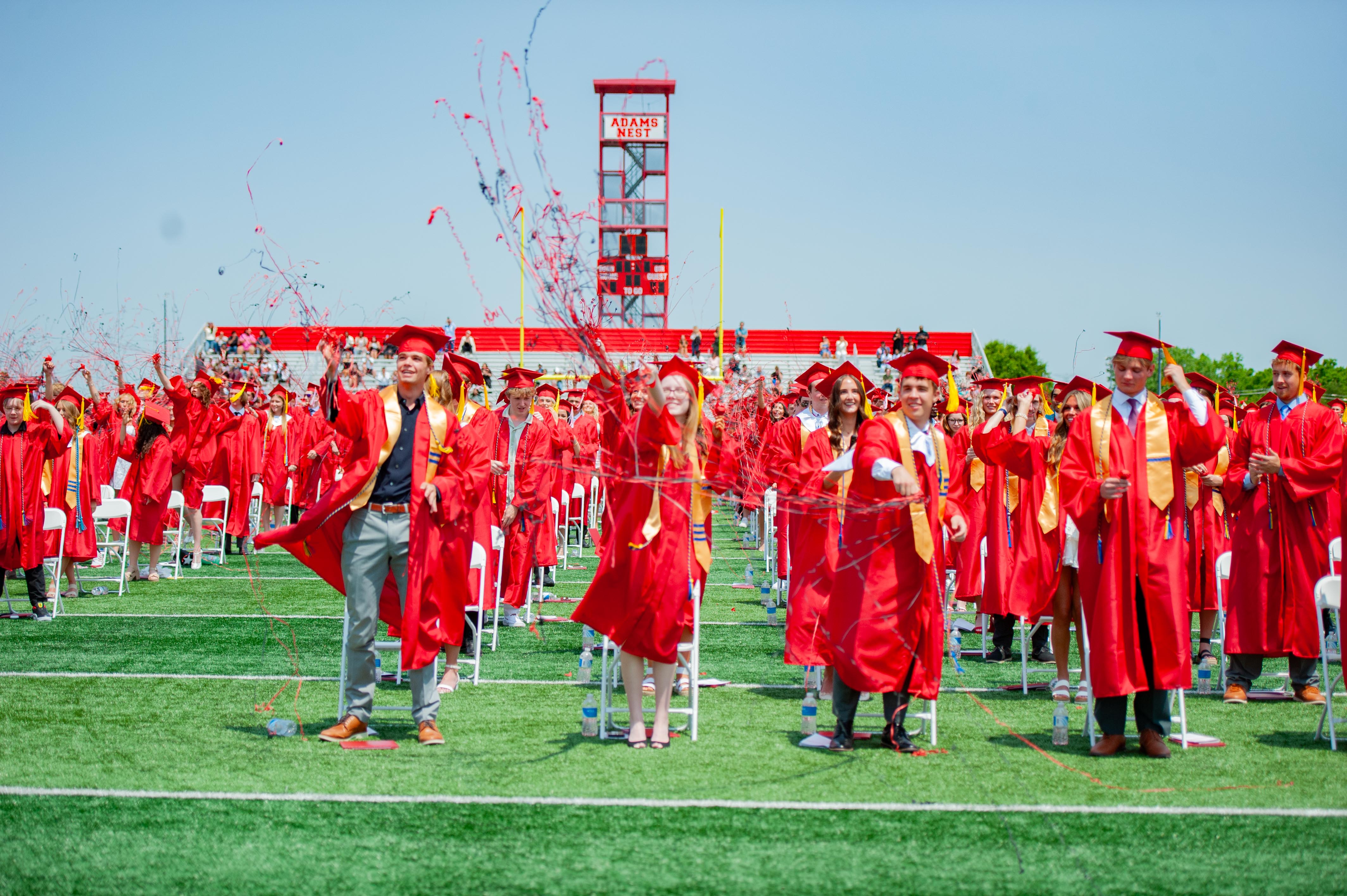 Graduates in red gowns celebrate with confetti on a green field, with a stadium scoreboard in the background.