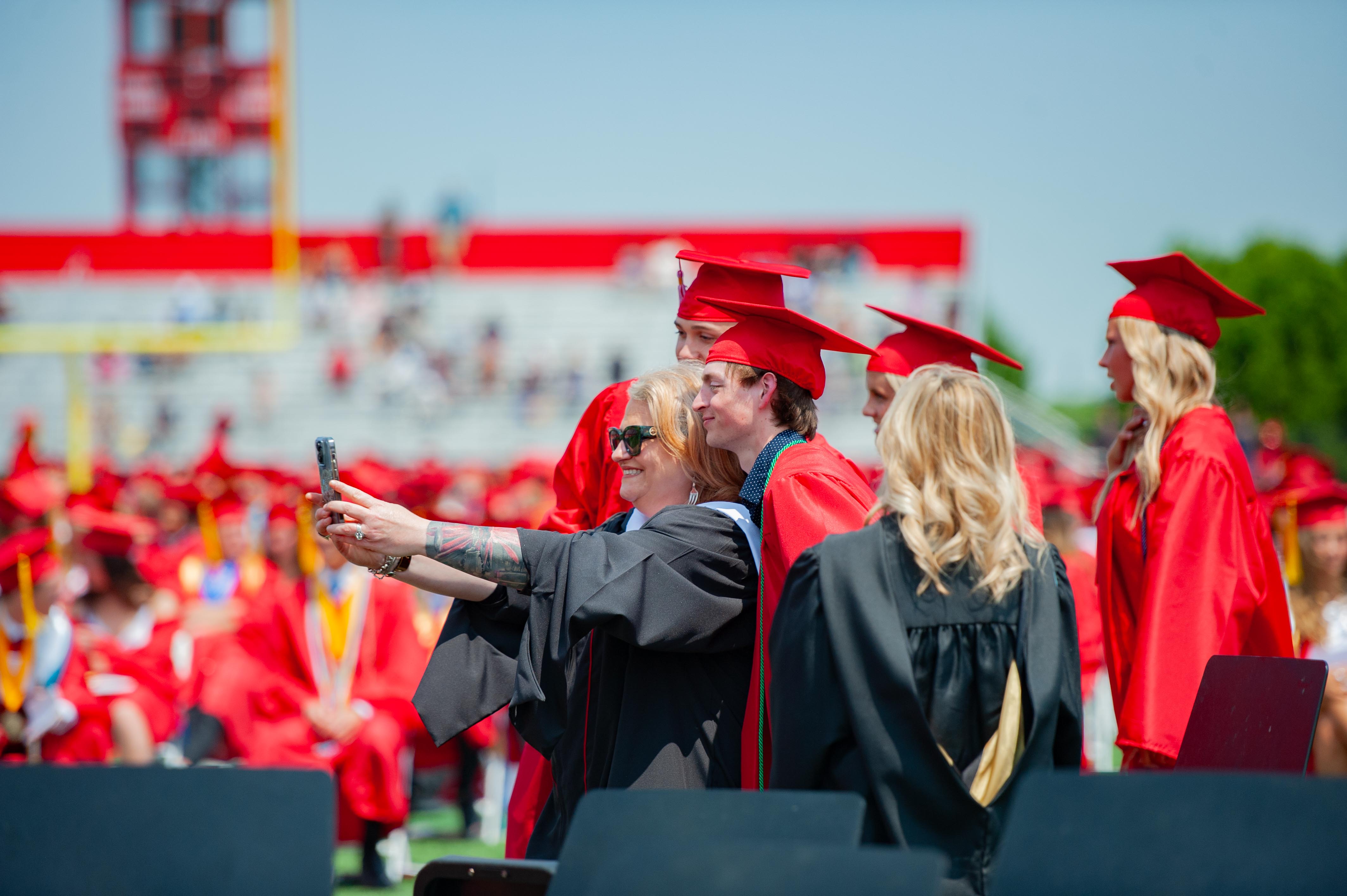 A group of graduates in red gowns and caps take a selfie in the foreground, with a stadium and cheering crowd in the background.