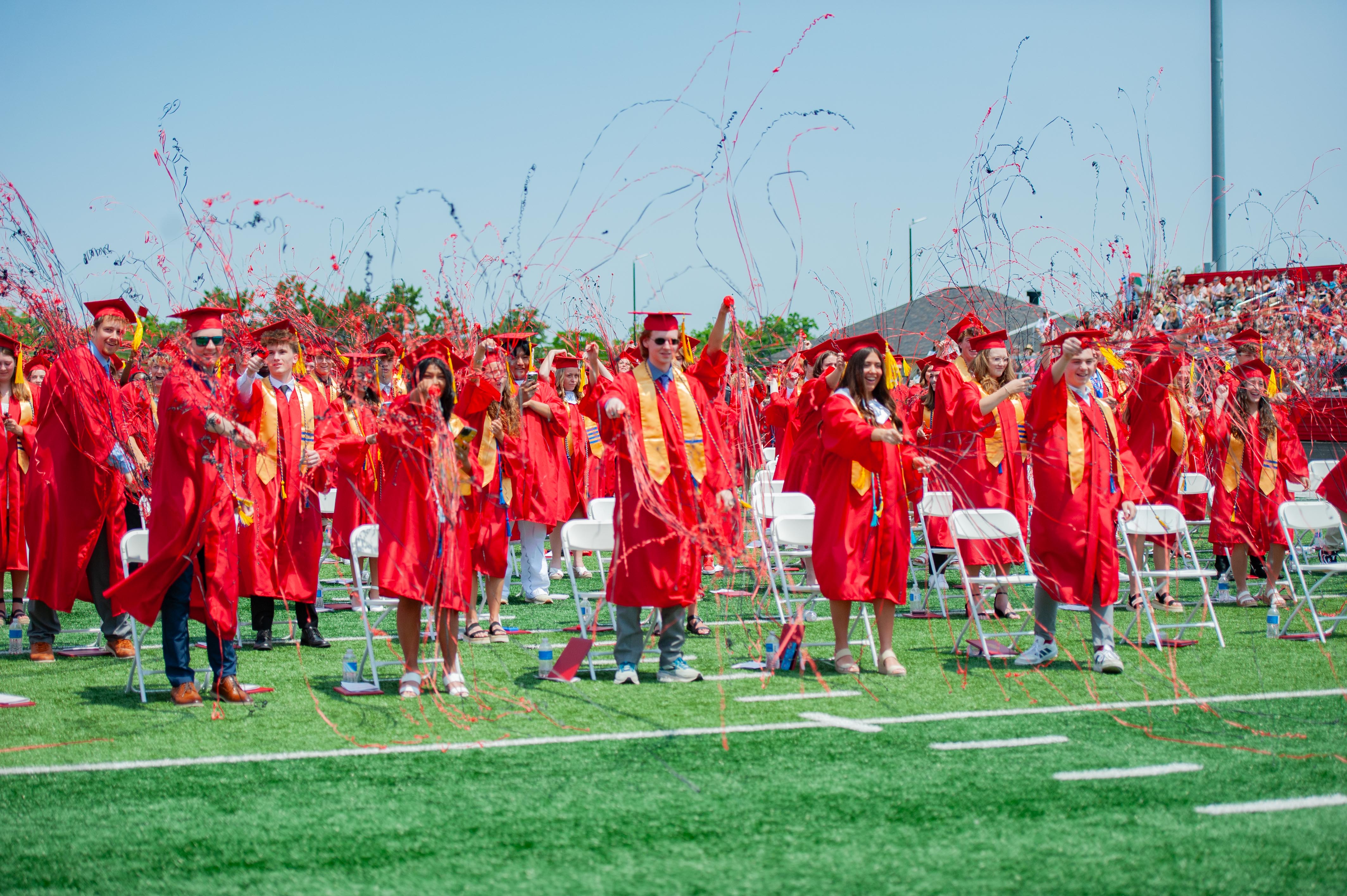 Graduates in red gowns and caps stand on a green field with confetti in the air, while a stadium filled with spectators is visible in the background.
