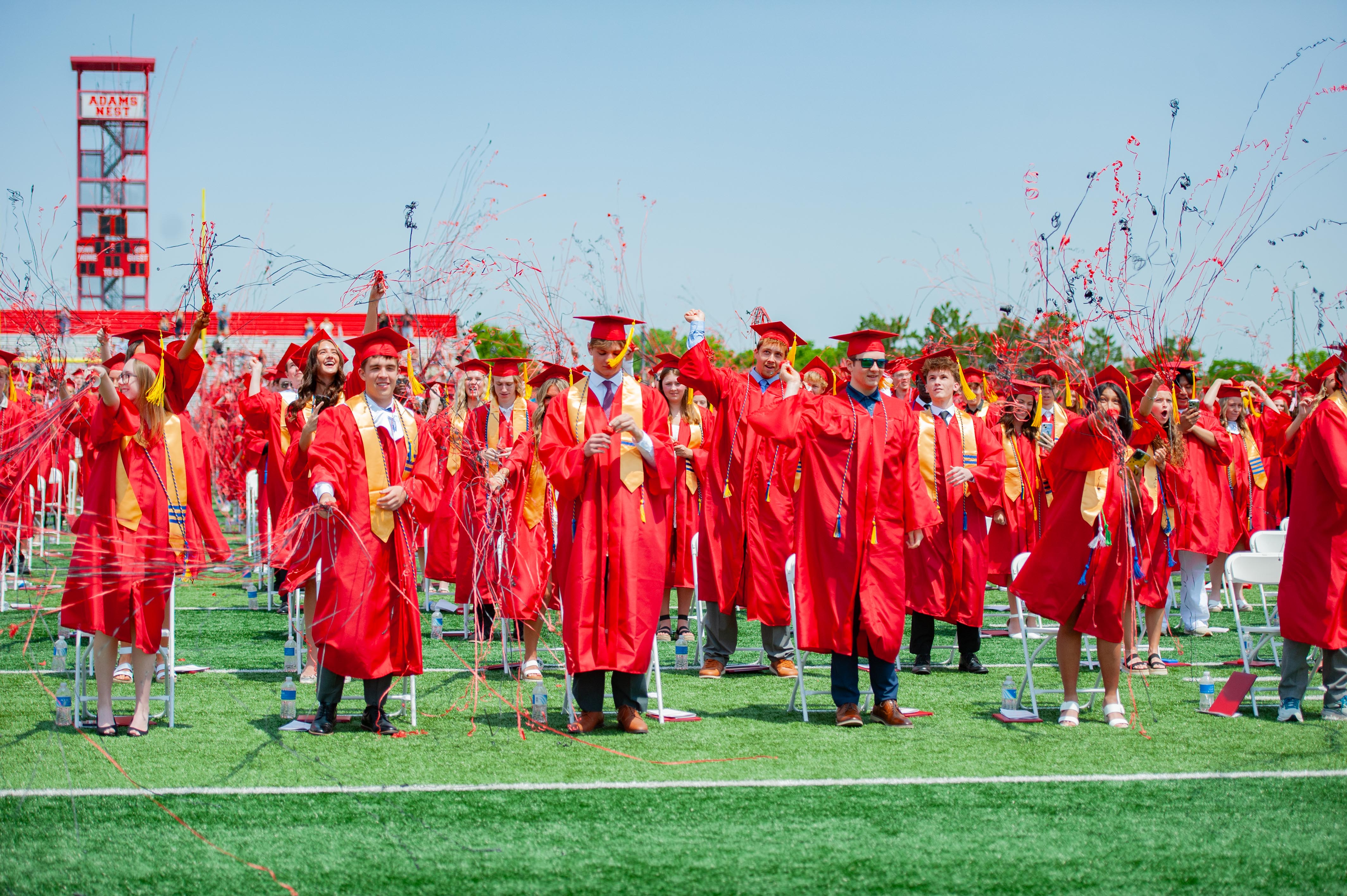 Graduates in red gowns celebrate with confetti on a green field in front of a stadium under a clear sky.