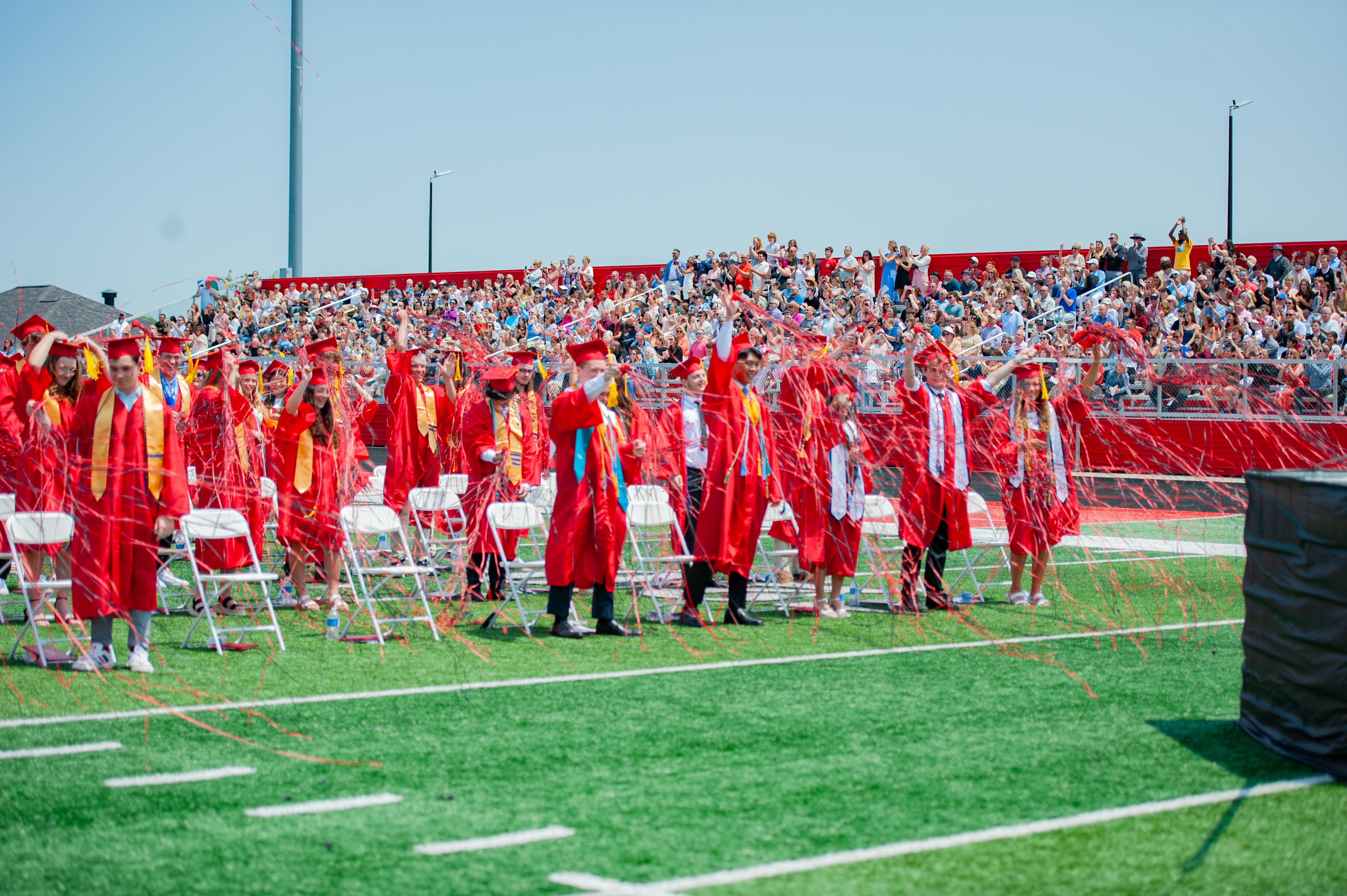 Graduates in red gowns celebrate with confetti on a green football field, while a large crowd watches from stadium bleachers in the background.