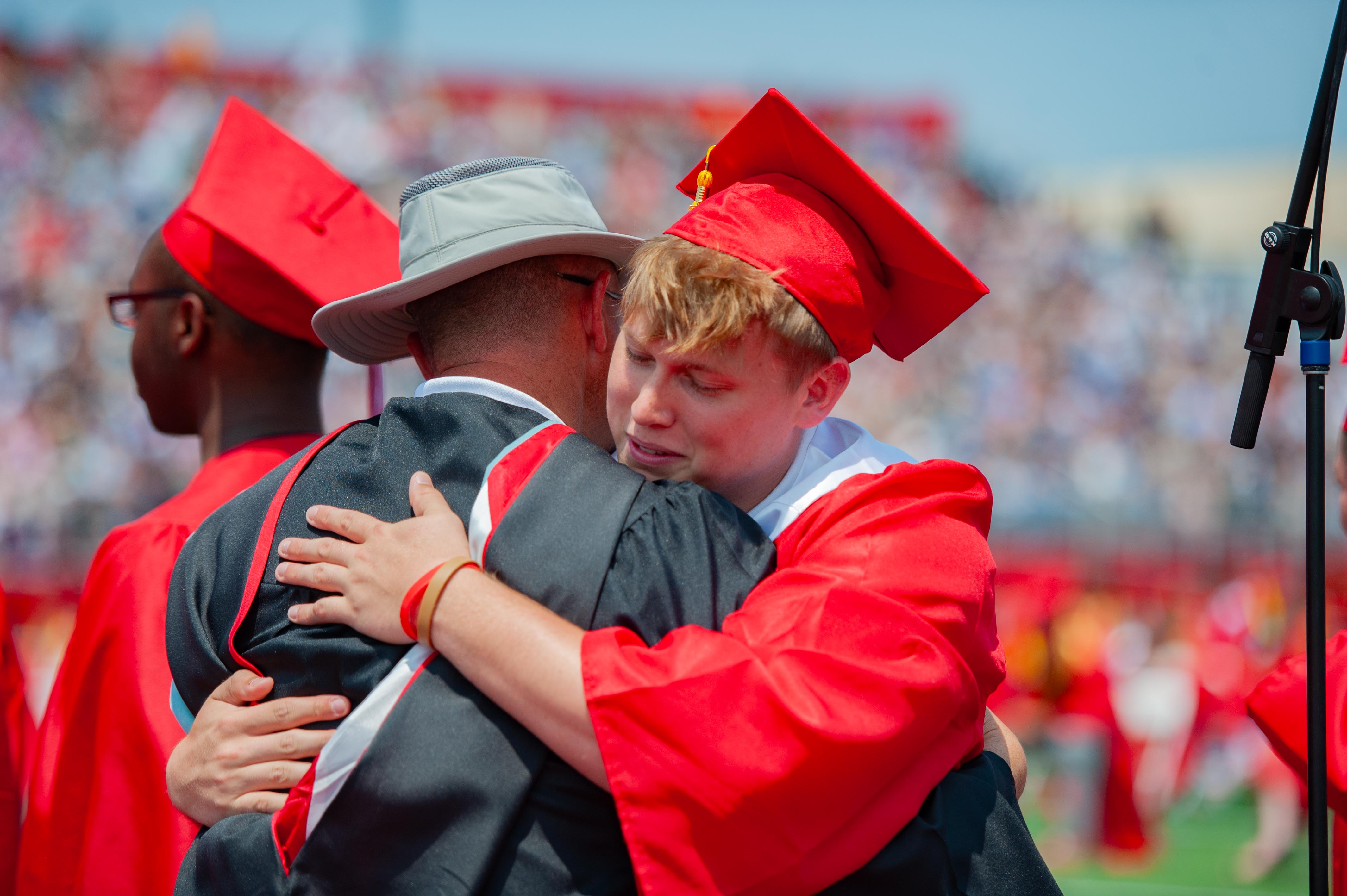 A young graduate in a red cap and gown tearfully hugs an adult in a hat and dark robe, with other graduates and a blurred stadium crowd in the background.