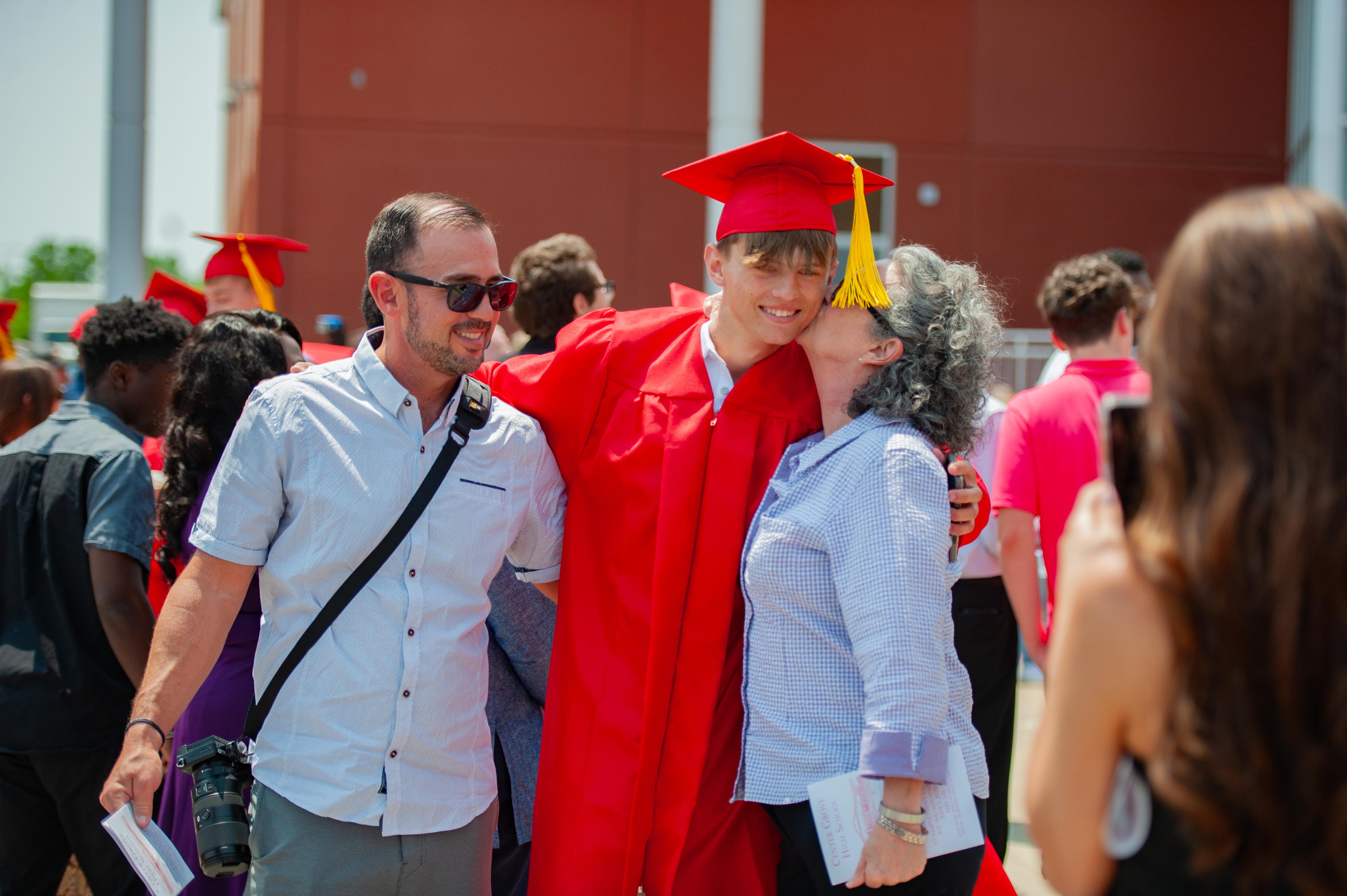 A graduating student in a red cap and gown is embraced by his parents, with other graduates and a building in the background.