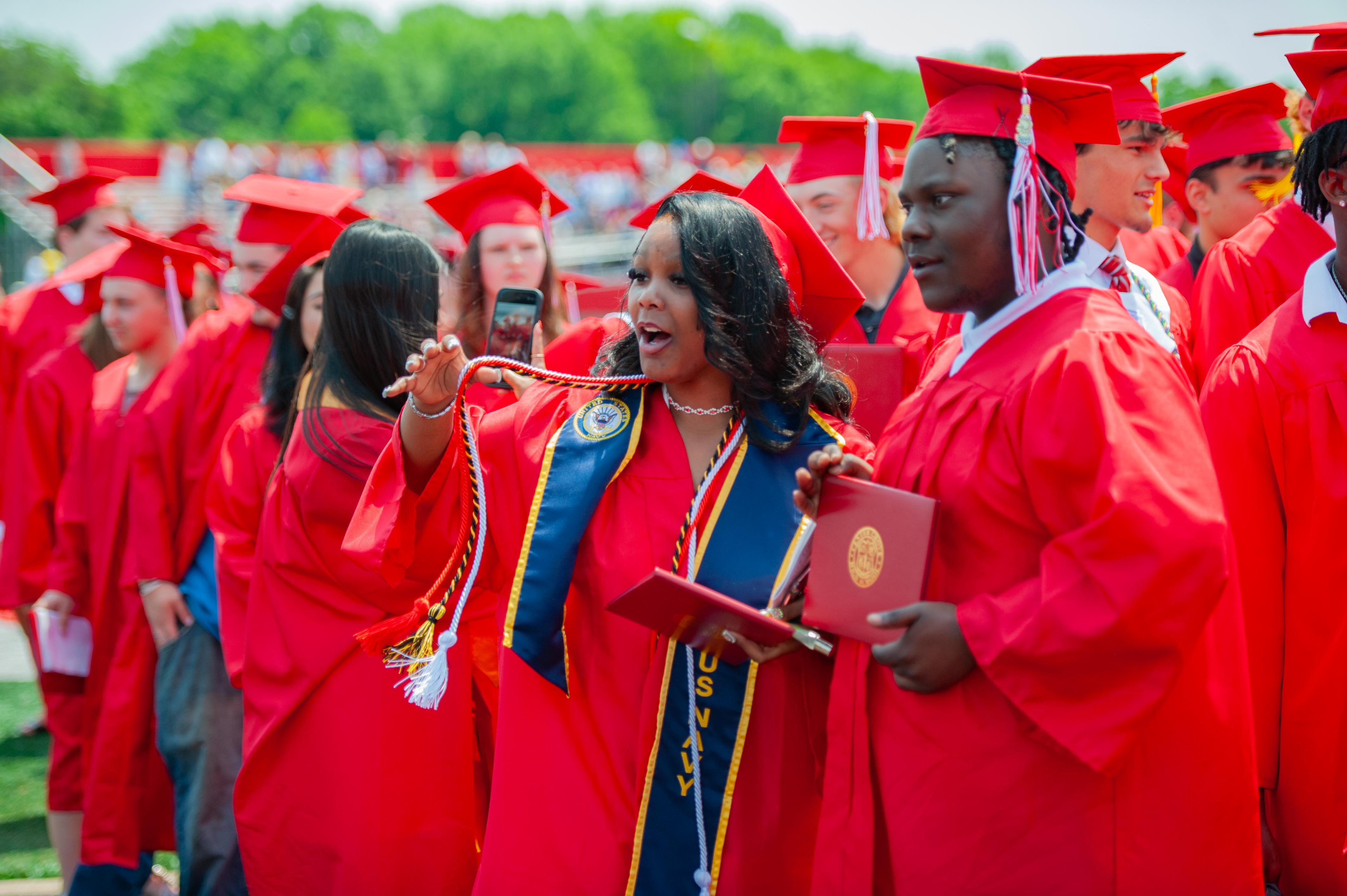 A group of graduates in red gowns and caps stand in a line, with a woman in the foreground holding a diploma and speaking, while a blurred background shows more graduates and trees.