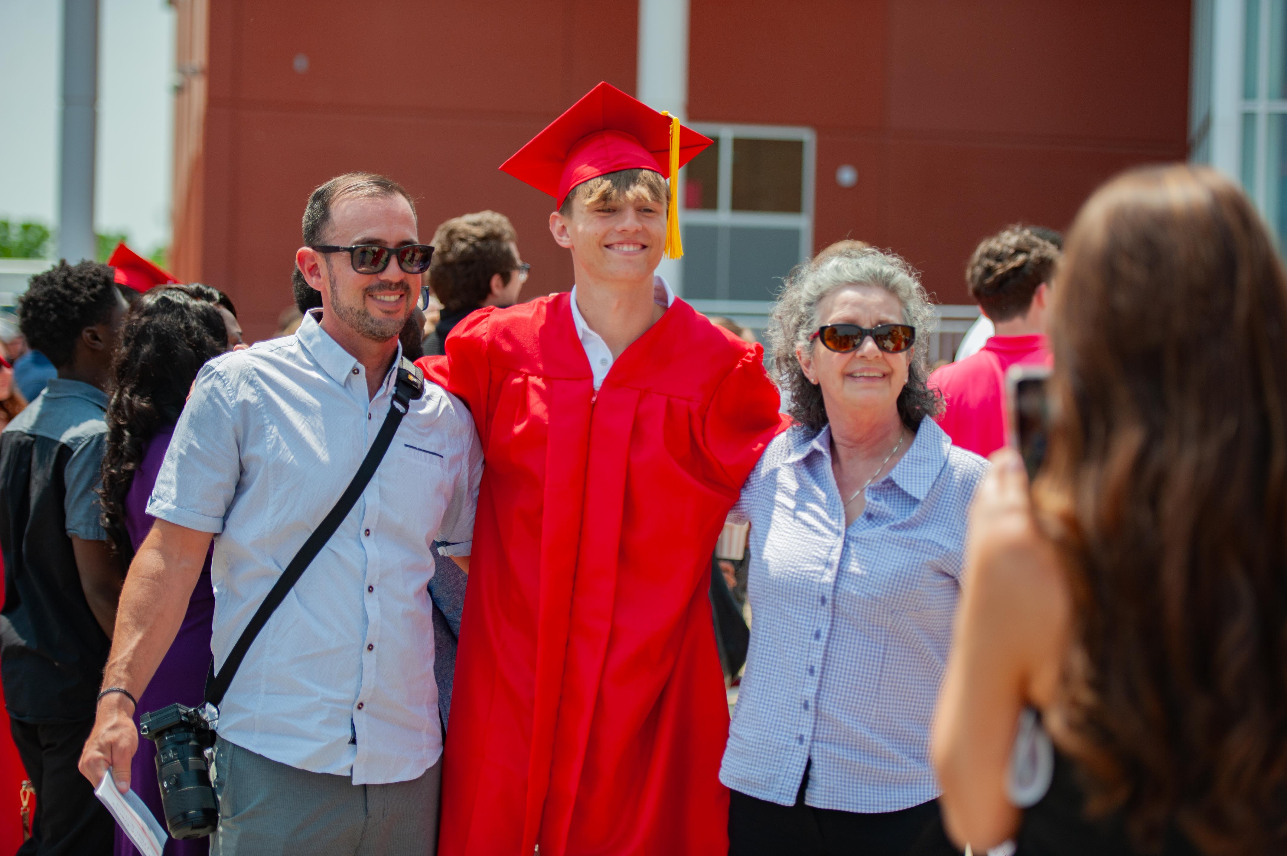 A graduating student in a red cap and gown poses with his parents in front of a building while another person takes a photo.