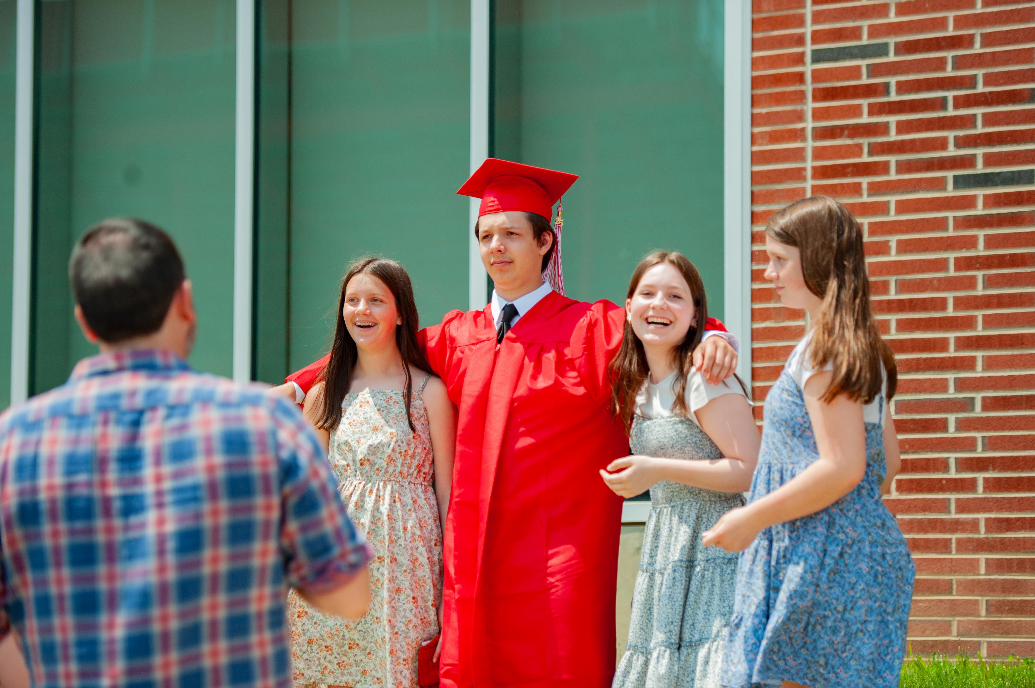 A graduating student in a red cap and gown poses with three smiling girls in front of a brick building with large windows.