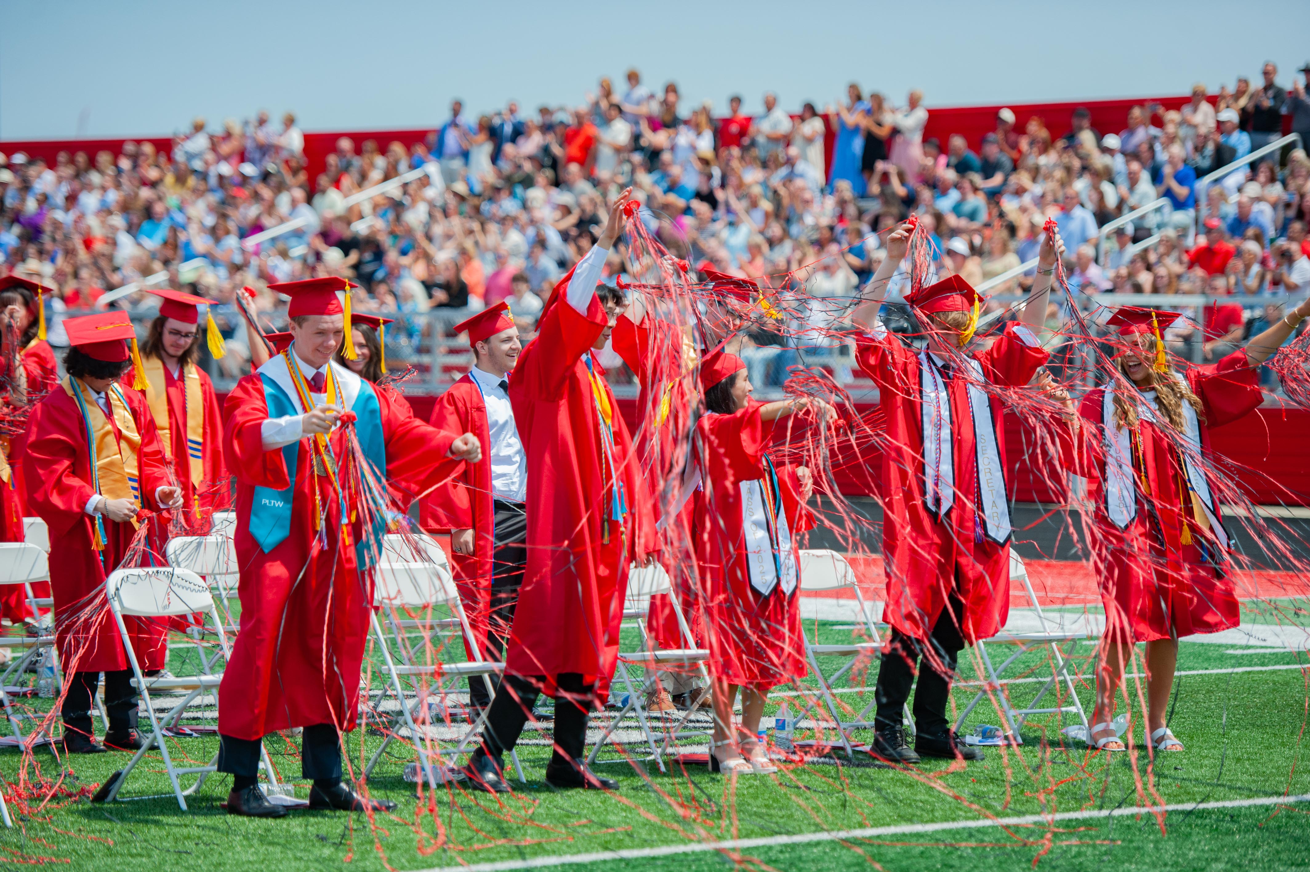 Graduates in red gowns celebrate with streamers on a football field in front of a cheering crowd in the stands.