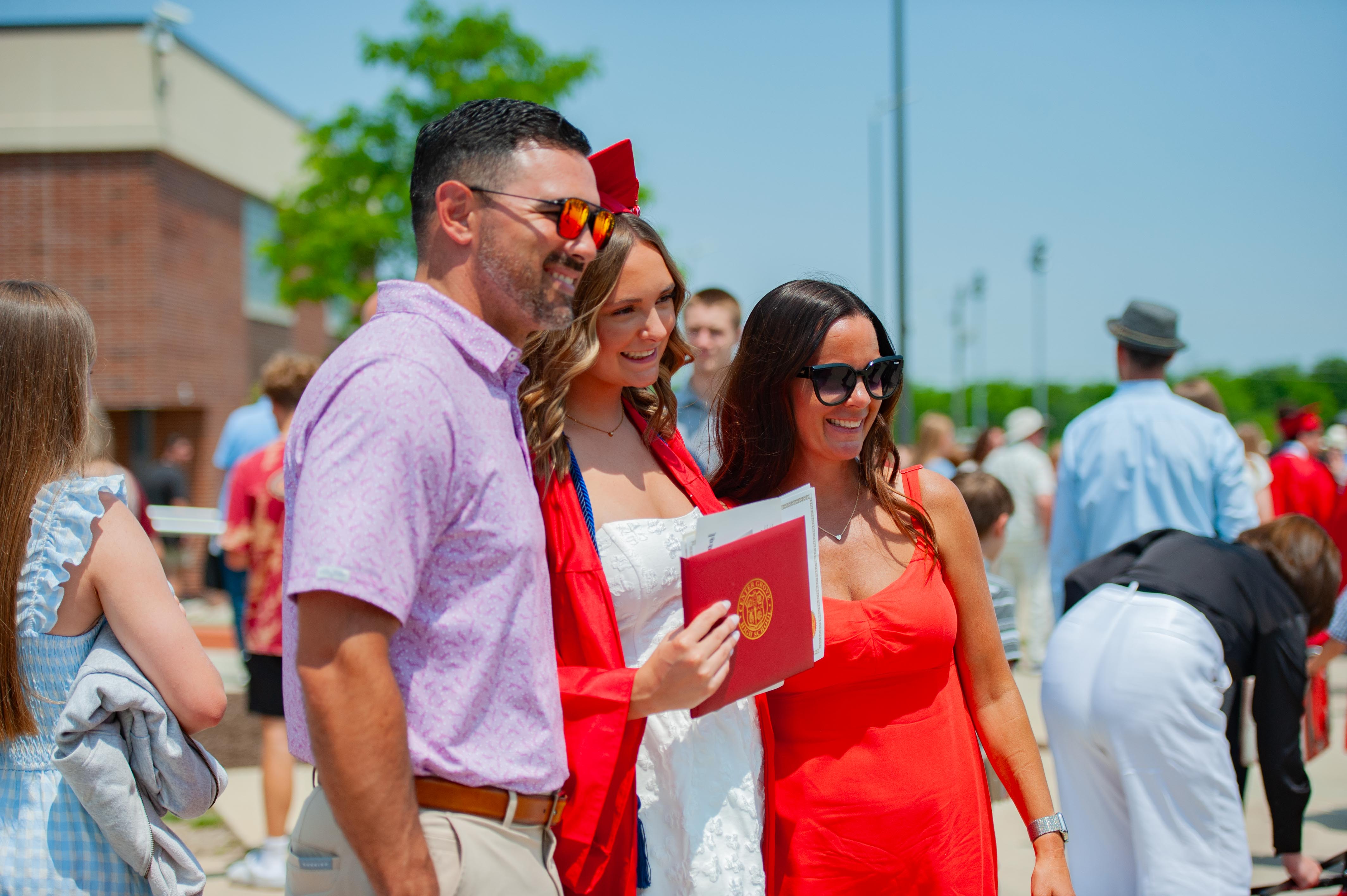 A smiling family poses for a photo in the foreground with a graduation diploma, while a crowd of people in casual and formal attire are visible in the blurred background under a bright sky.