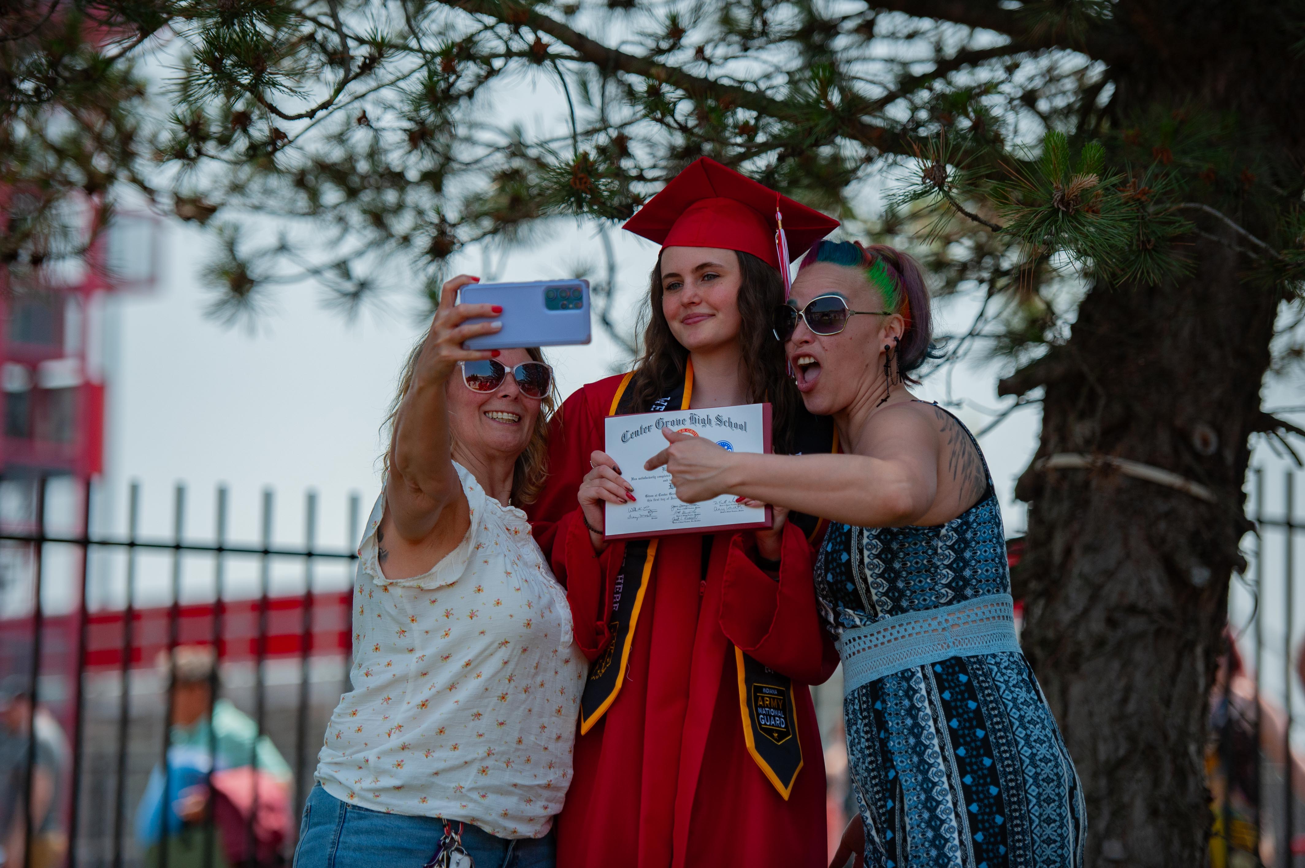 A graduate in a red cap and gown poses for a selfie with two women under a tree, with a red building and fence in the blurred background.