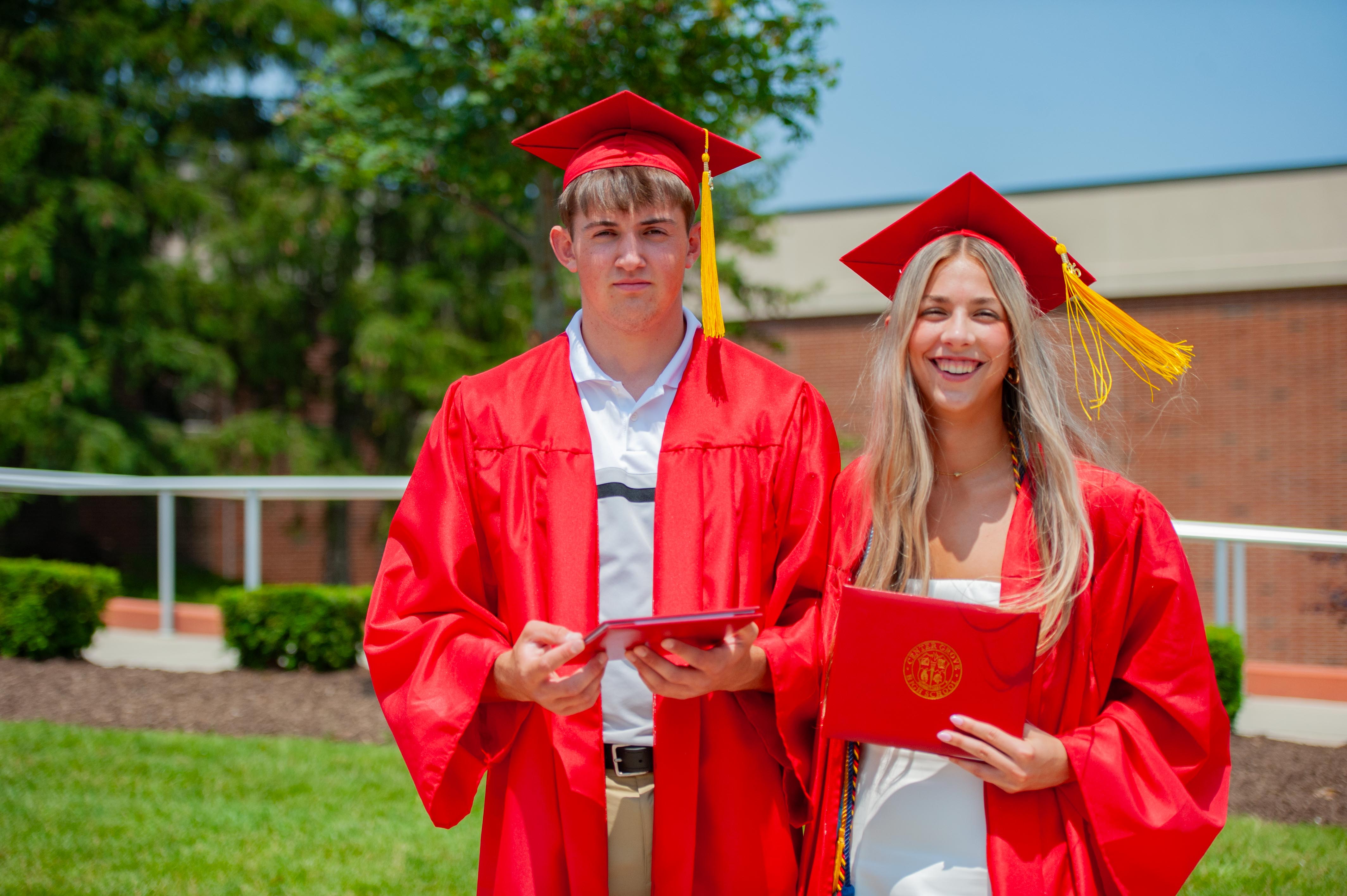 Two graduates in red caps and gowns hold diplomas in front of a brick building and green trees.