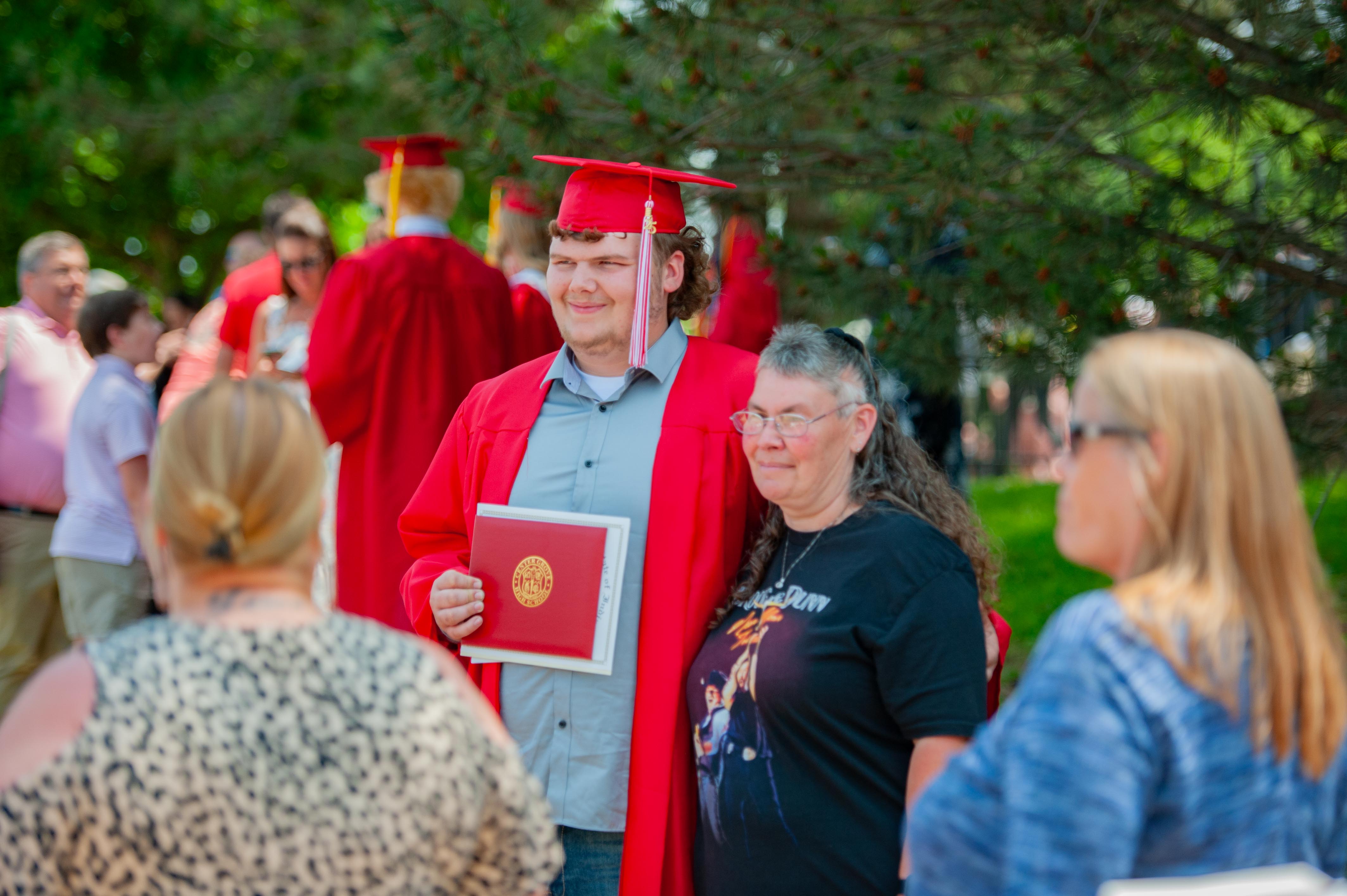 A young man in a graduation cap and gown holds his diploma while standing with a woman, as other graduates and onlookers are visible in the blurred background.