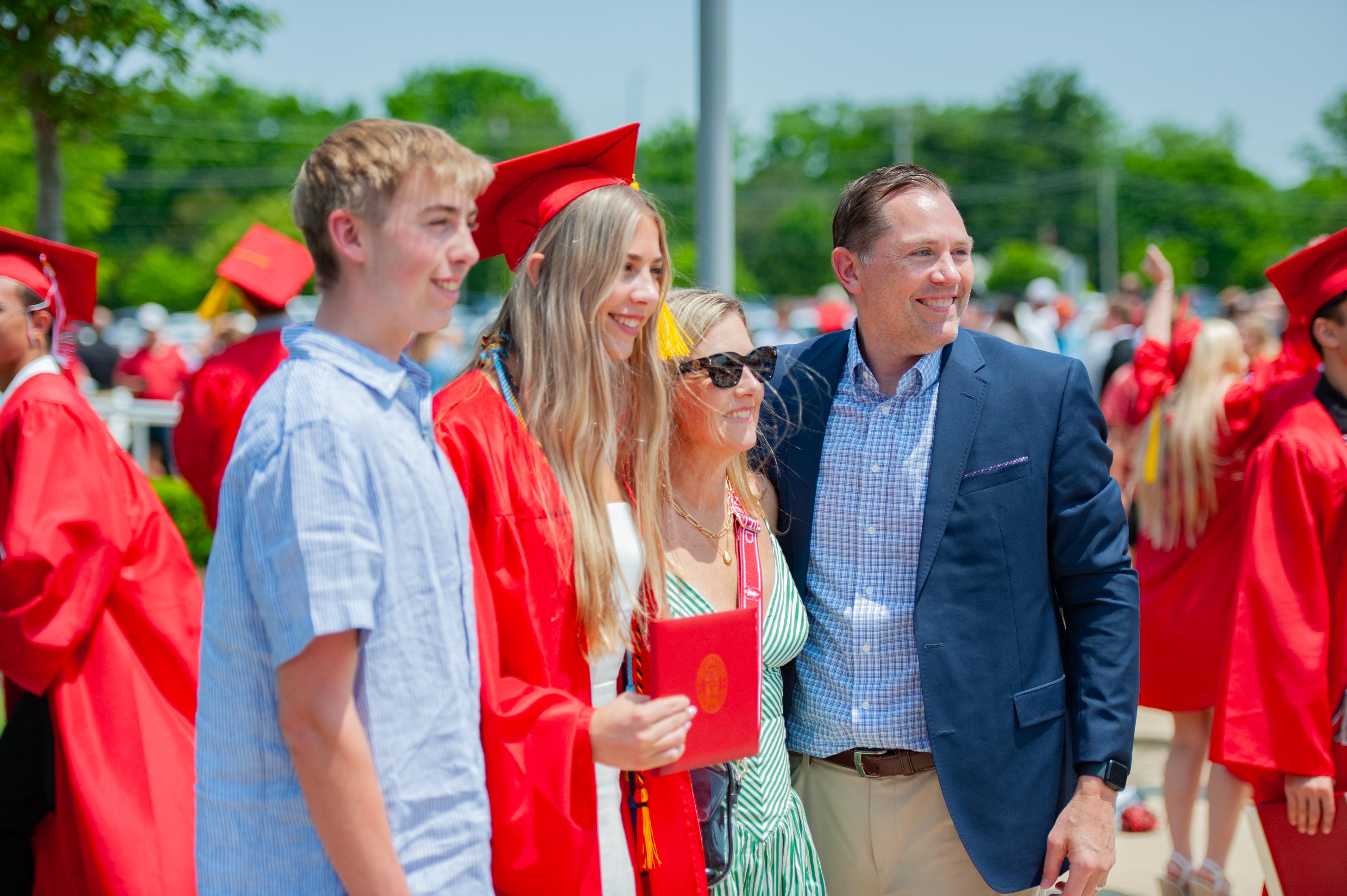 A family poses for a photo in the foreground with other graduates in red gowns and caps in the blurred background.