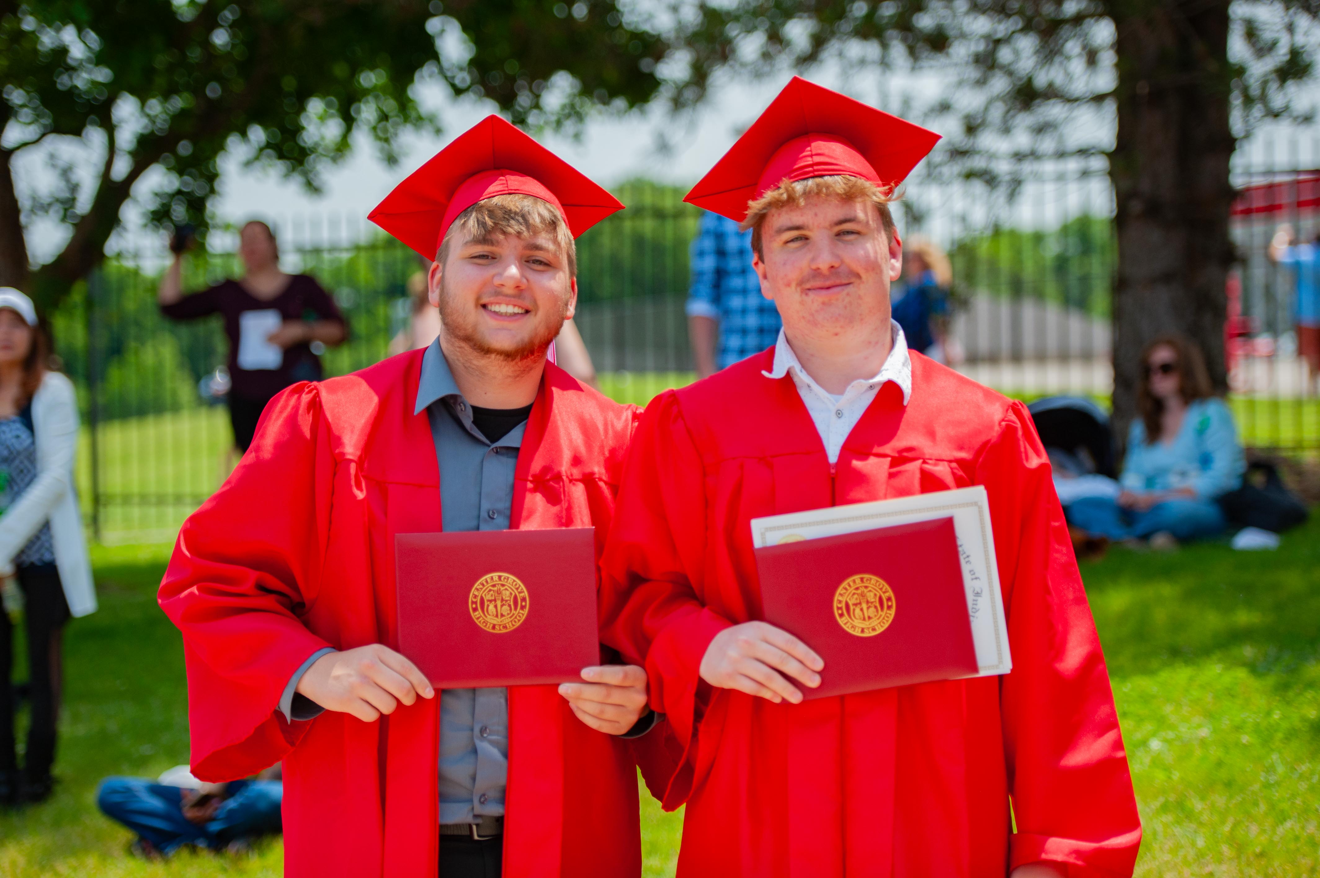 Two smiling graduates in red caps and gowns hold diplomas in the foreground, with blurred figures and trees in the background.