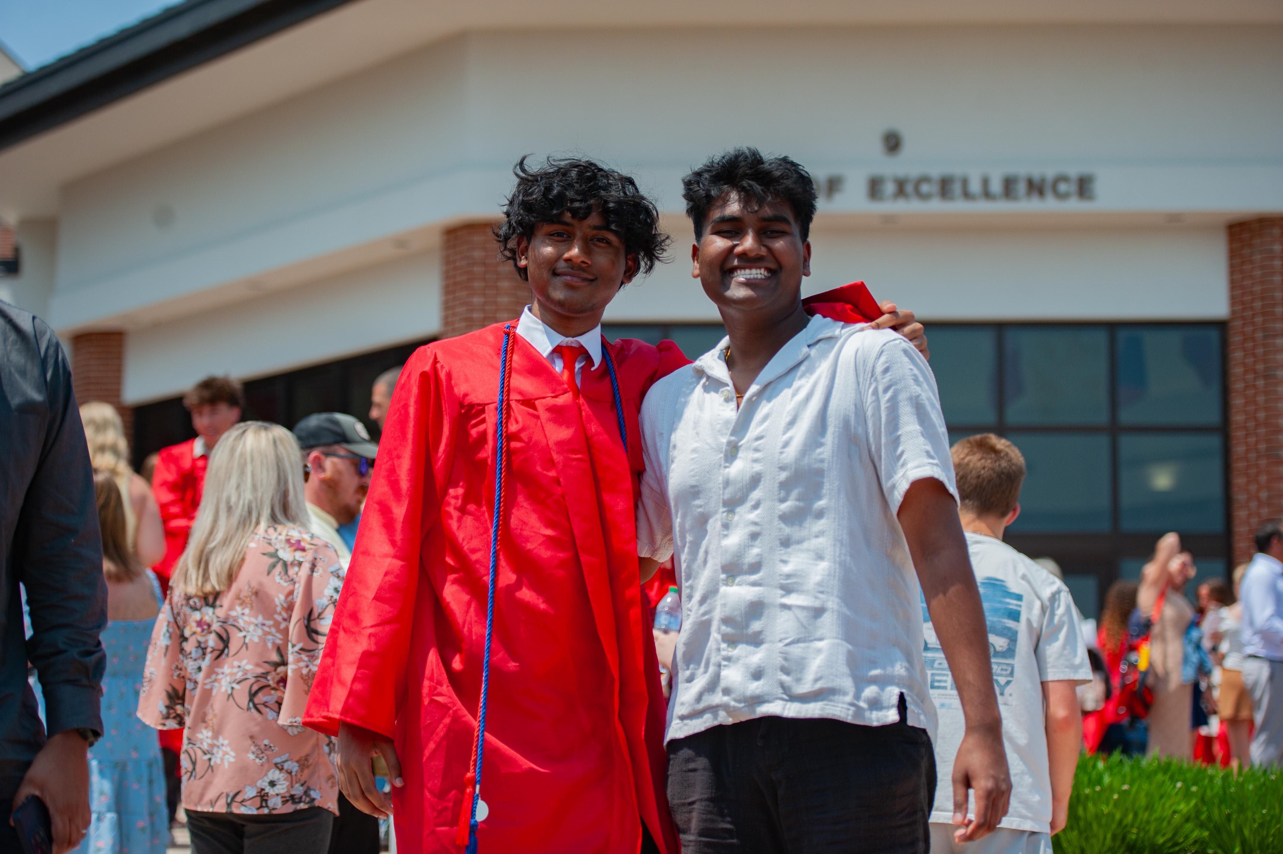 Two young men, one in a red graduation gown and the other in a white shirt, pose for a photo with a building labeled "OF EXCELLENCE" in the background.