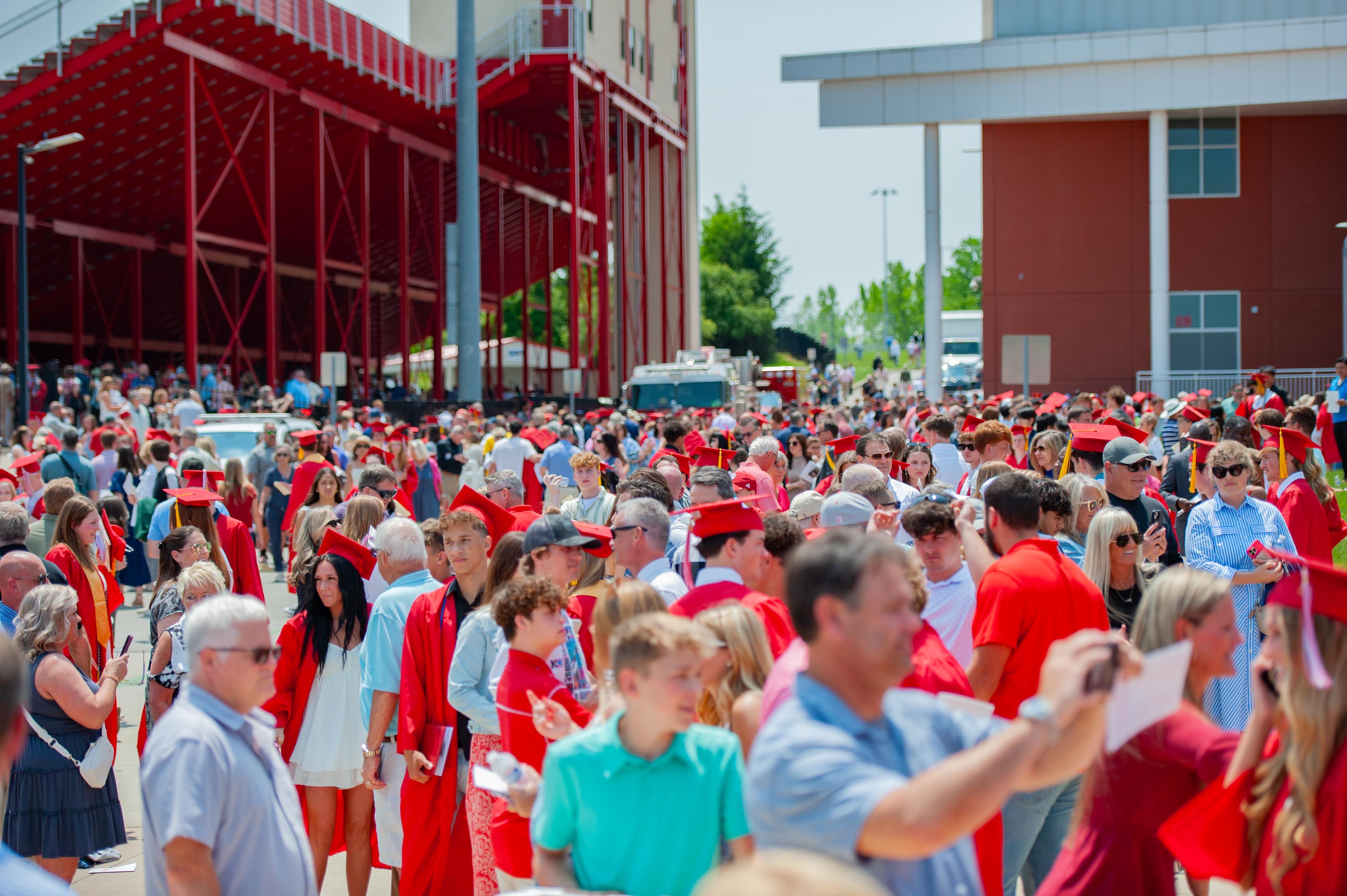 A large crowd of graduates in red gowns and caps fill the foreground, with a stadium and modern buildings in the background.