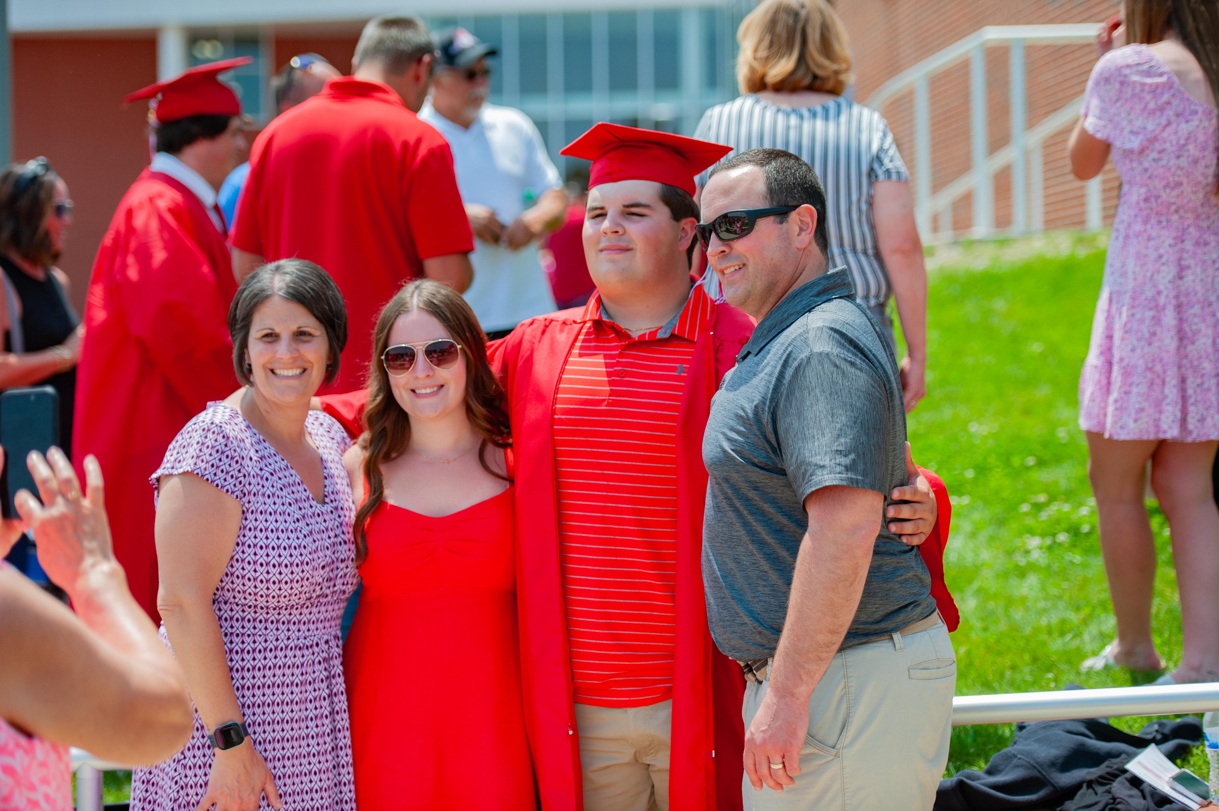 A family poses for a photo in front of a crowd of graduates in red caps and gowns.