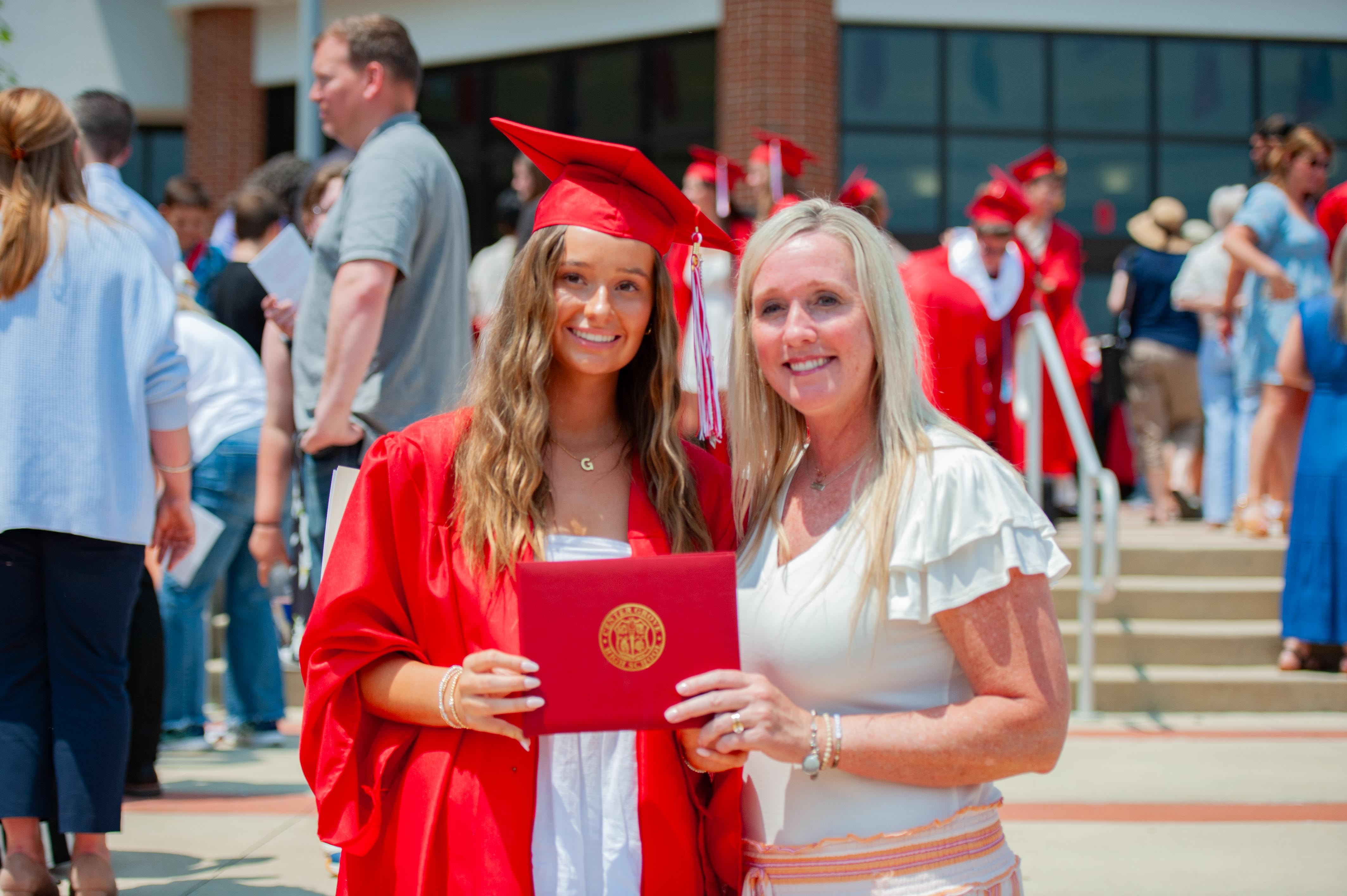 A smiling graduate in a red cap and gown holds a diploma with her mother in front of a crowd of people at a graduation ceremony.