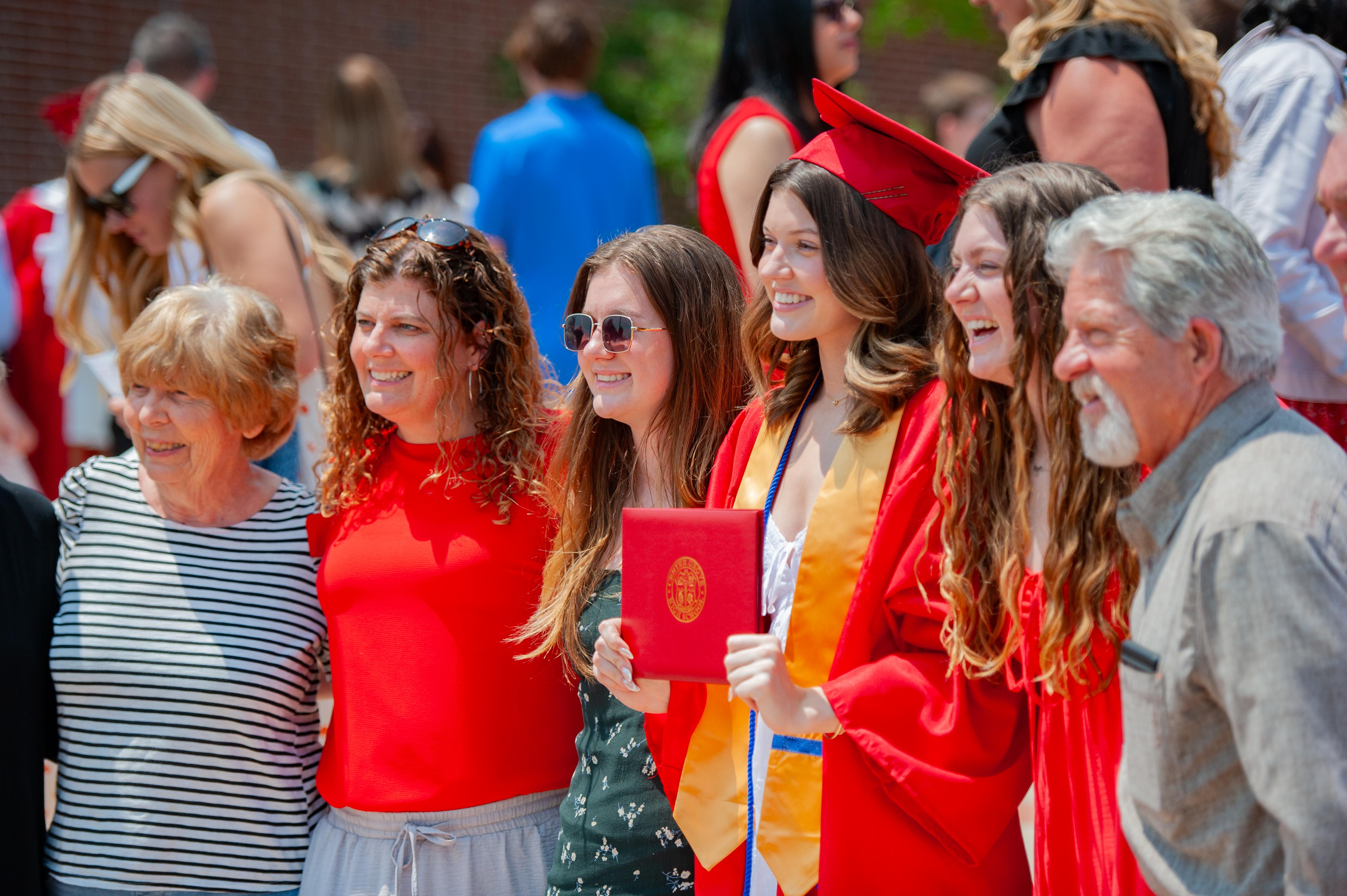 A group of smiling people, some in graduation gowns, pose for a photo with a brick building in the blurred background.
